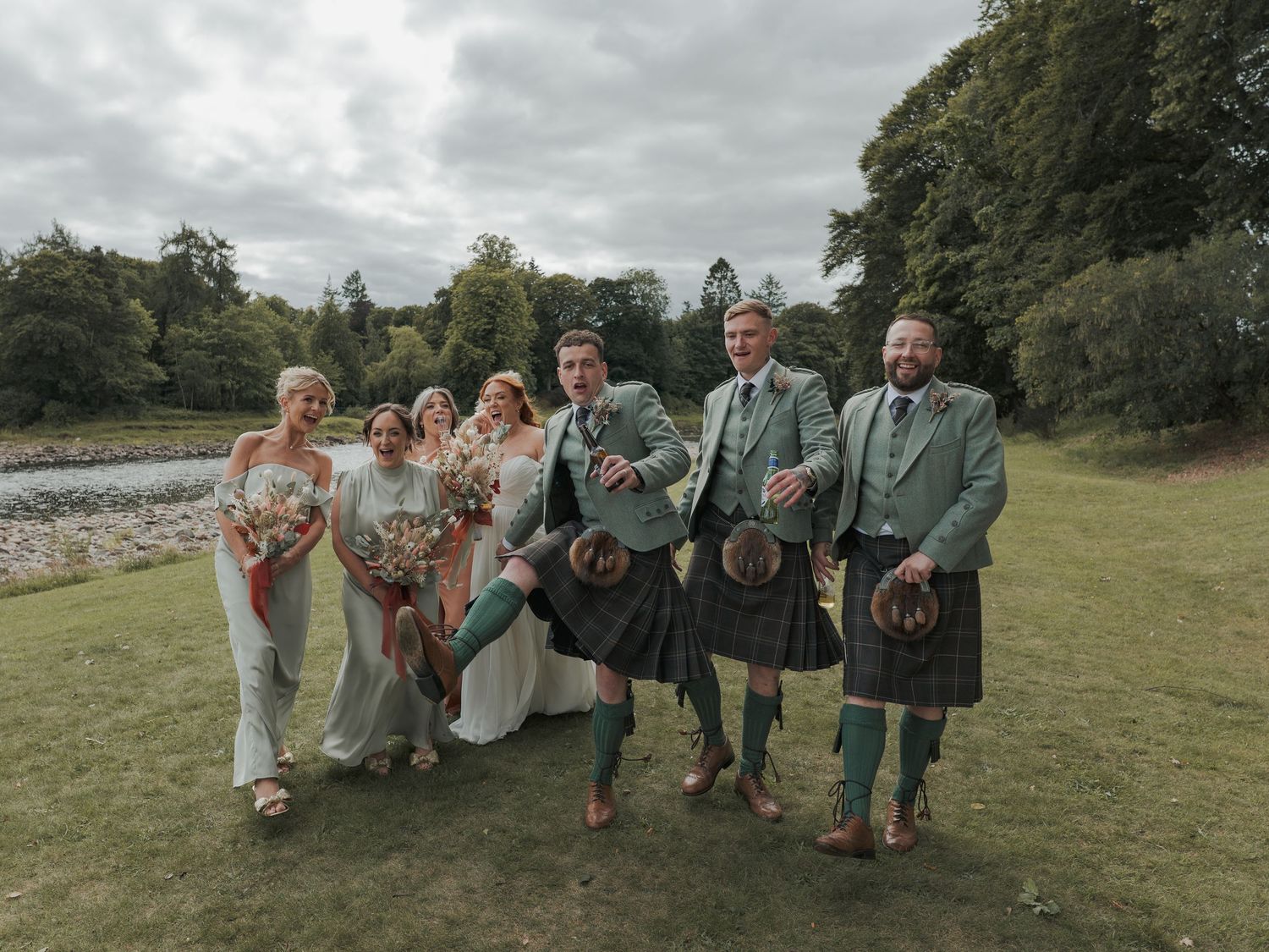 Wedding party poses playfully in matching formal attire on grassy hillside during outdoor celebration.