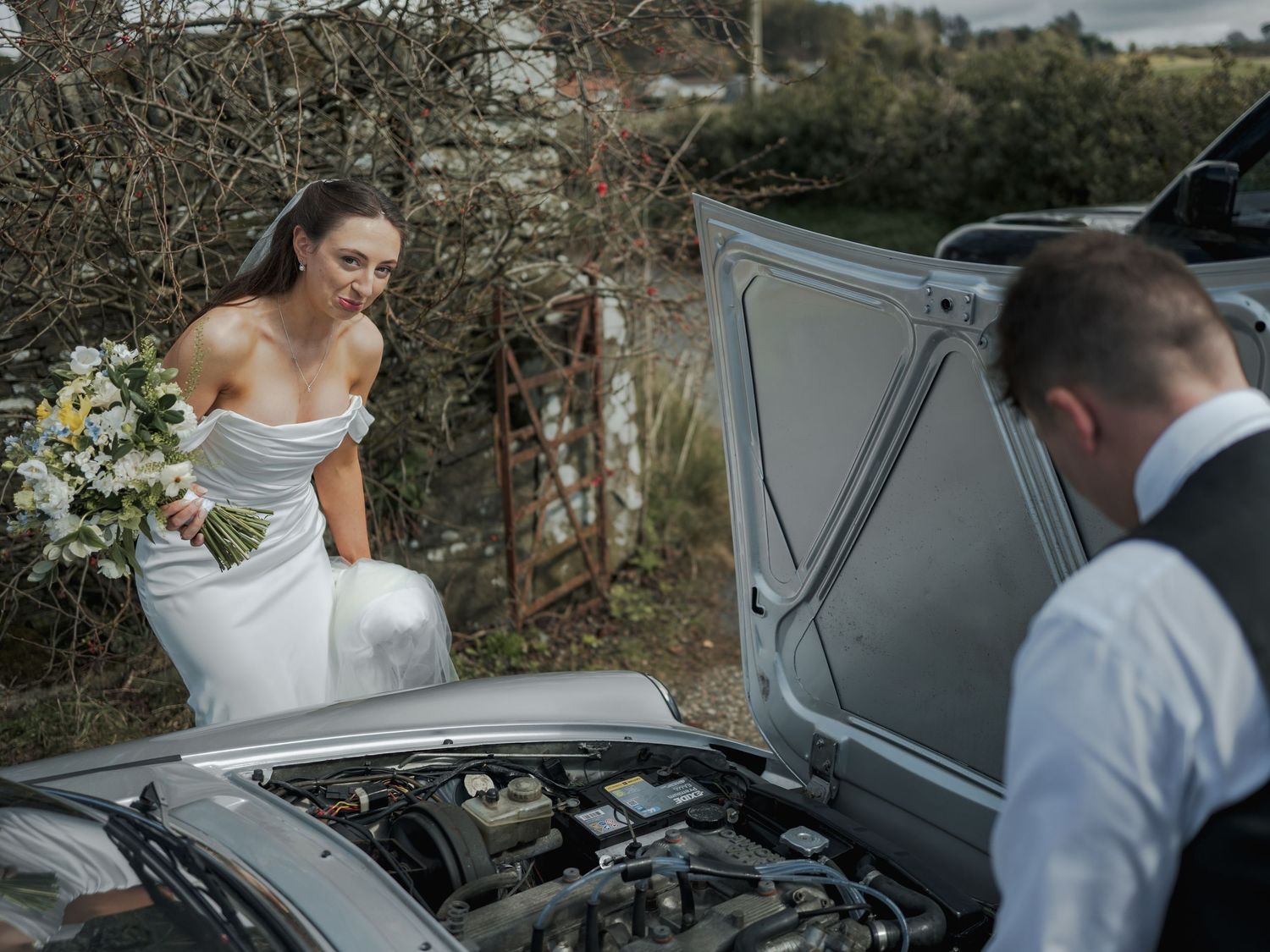 Bride in white gown holds bouquet while groom checks car engine on wedding day.