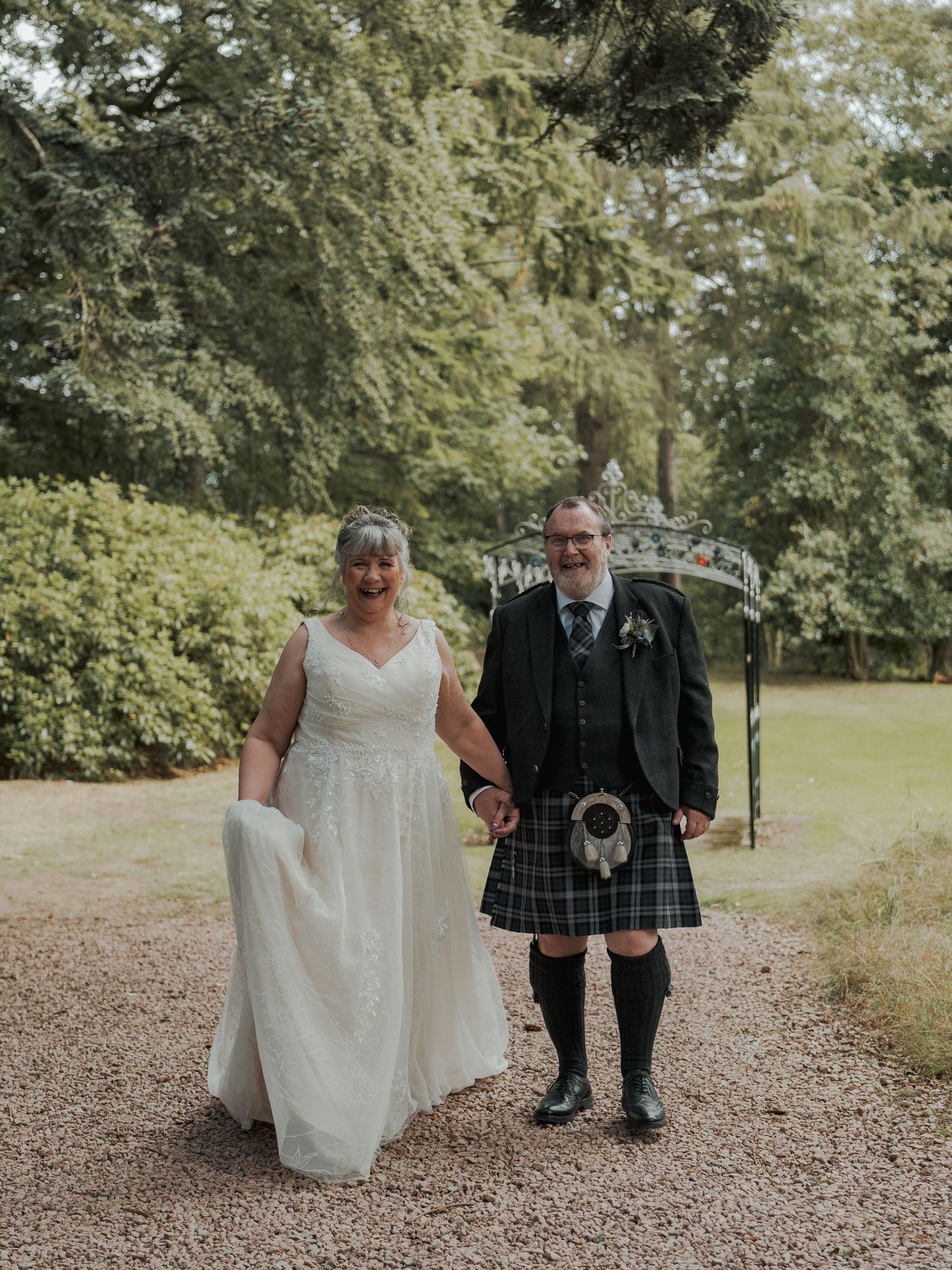 Bride in white gown and groom in traditional Scottish kilt walk hand in hand on a gravel path surrounded by lush greenery.