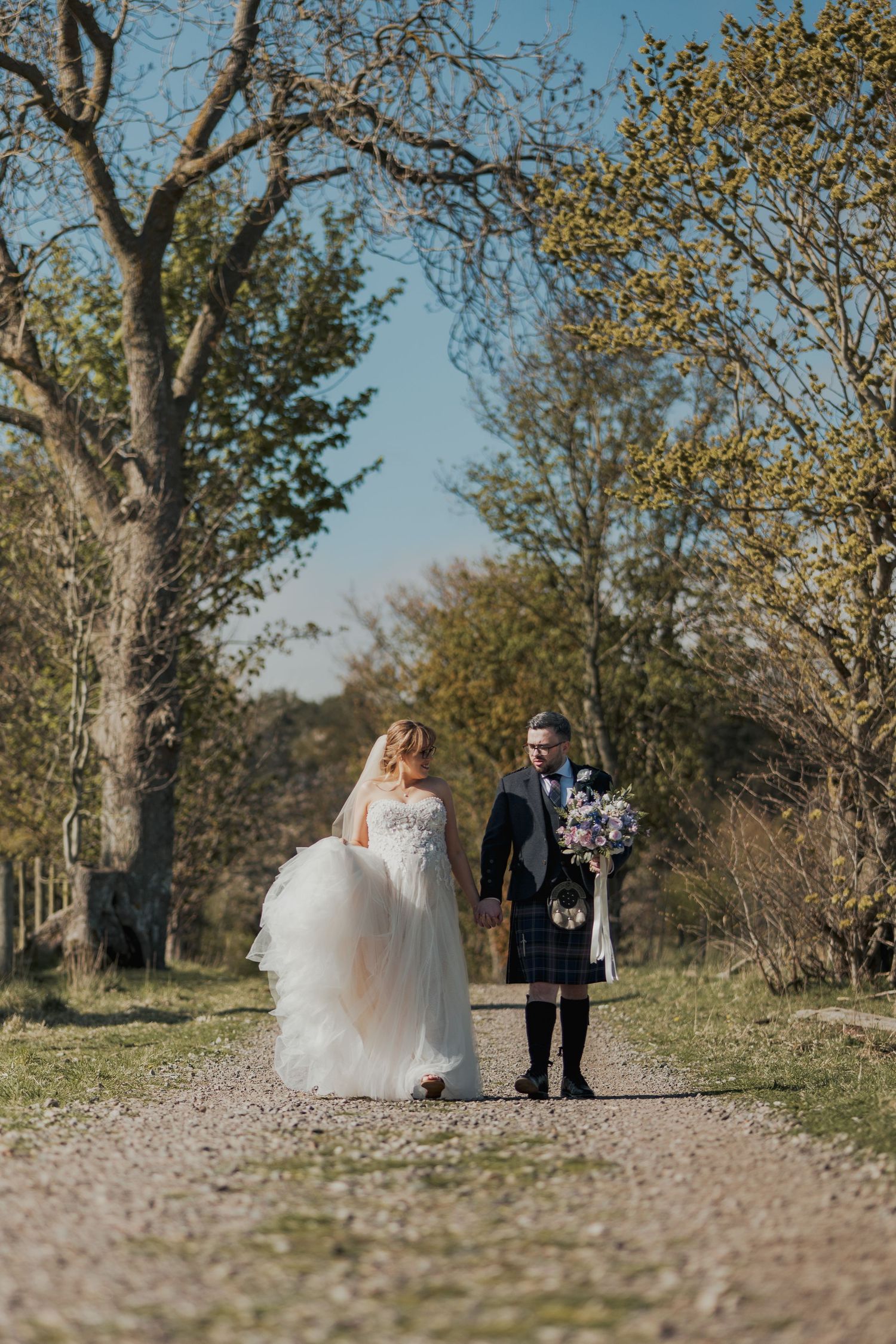 Bride in flowing white gown and kilted groom walk hand-in-hand along sunlit gravel path surrounded by autumn trees.