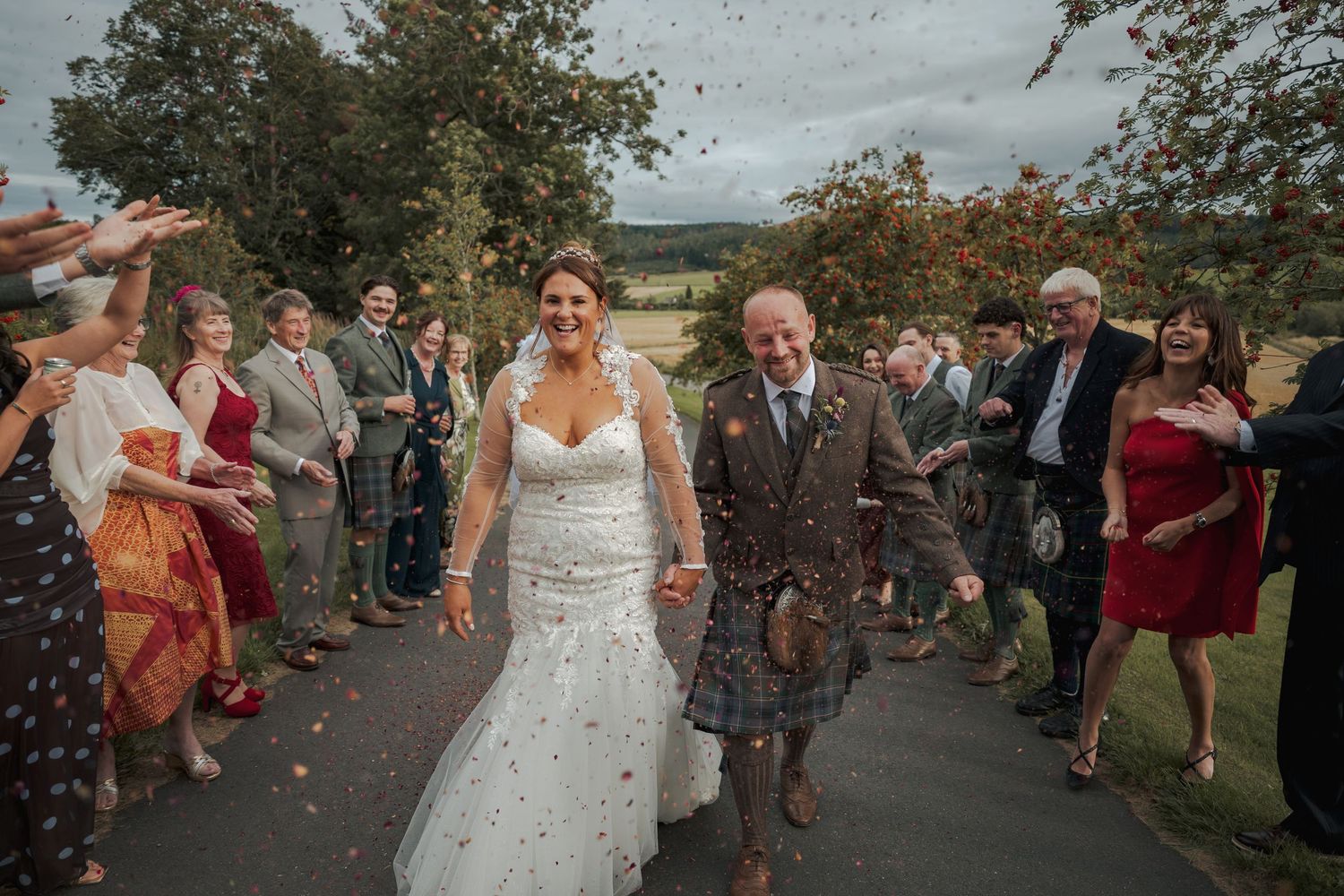 Bride and groom walk through confetti shower with smiling wedding guests lining path at outdoor Scottish countryside wedding.