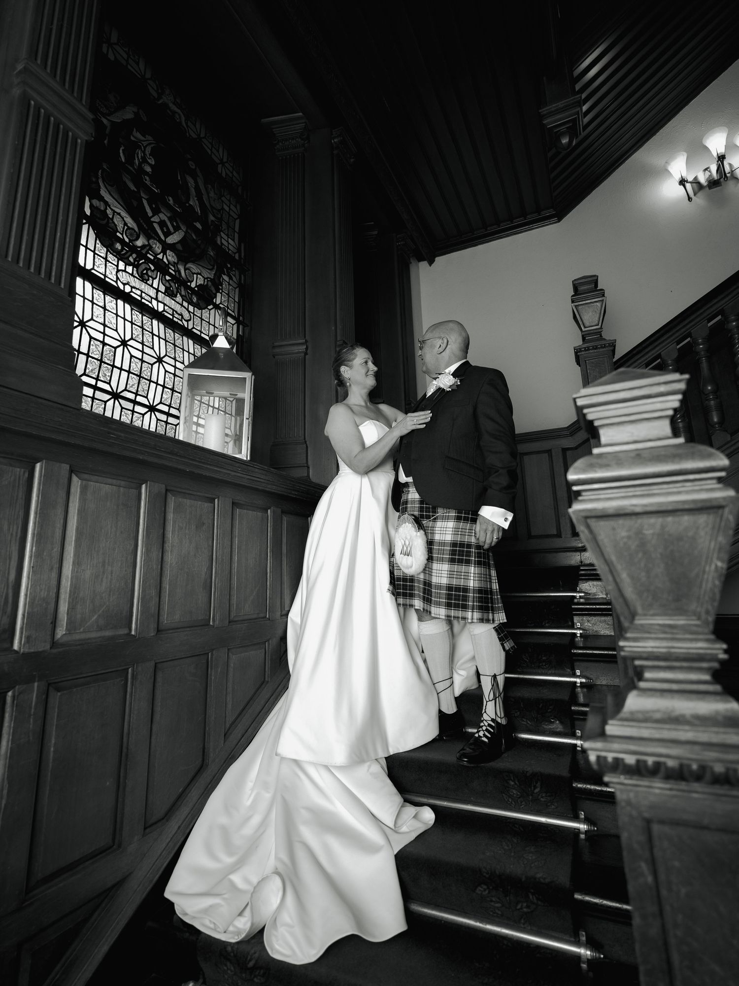 Black and white wedding photo of bride in white gown and groom in tartan kilt on grand wooden staircase.