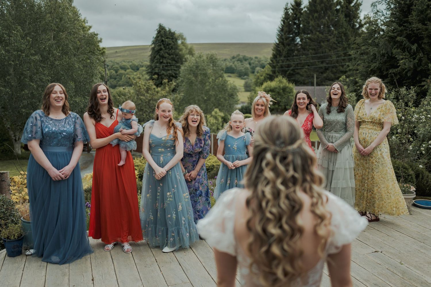 Bride faces colorful bridesmaids in floral gowns on a wooden deck surrounded by lush green hills.