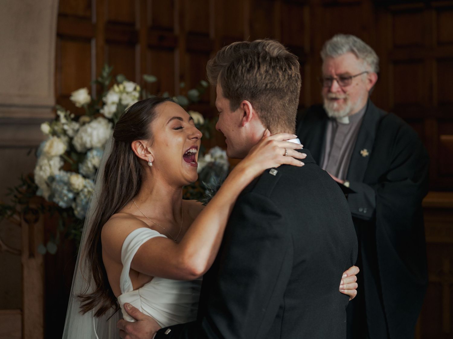 Bride and groom share first kiss at wedding ceremony with officiant standing behind them in elegant wood-paneled venue.
