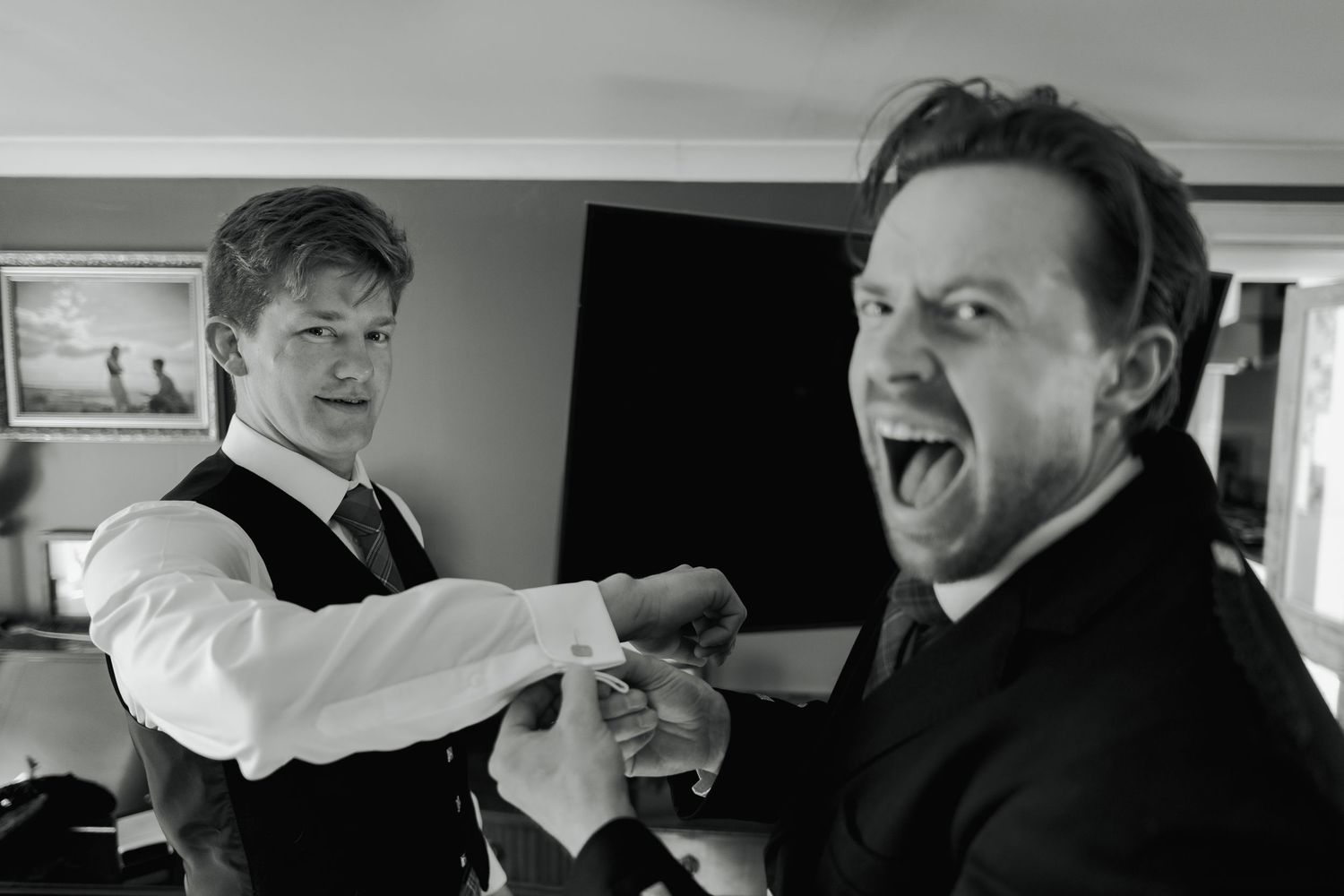 Black and white photo of two men in suits laughing and adjusting cufflinks at a wedding preparation.
