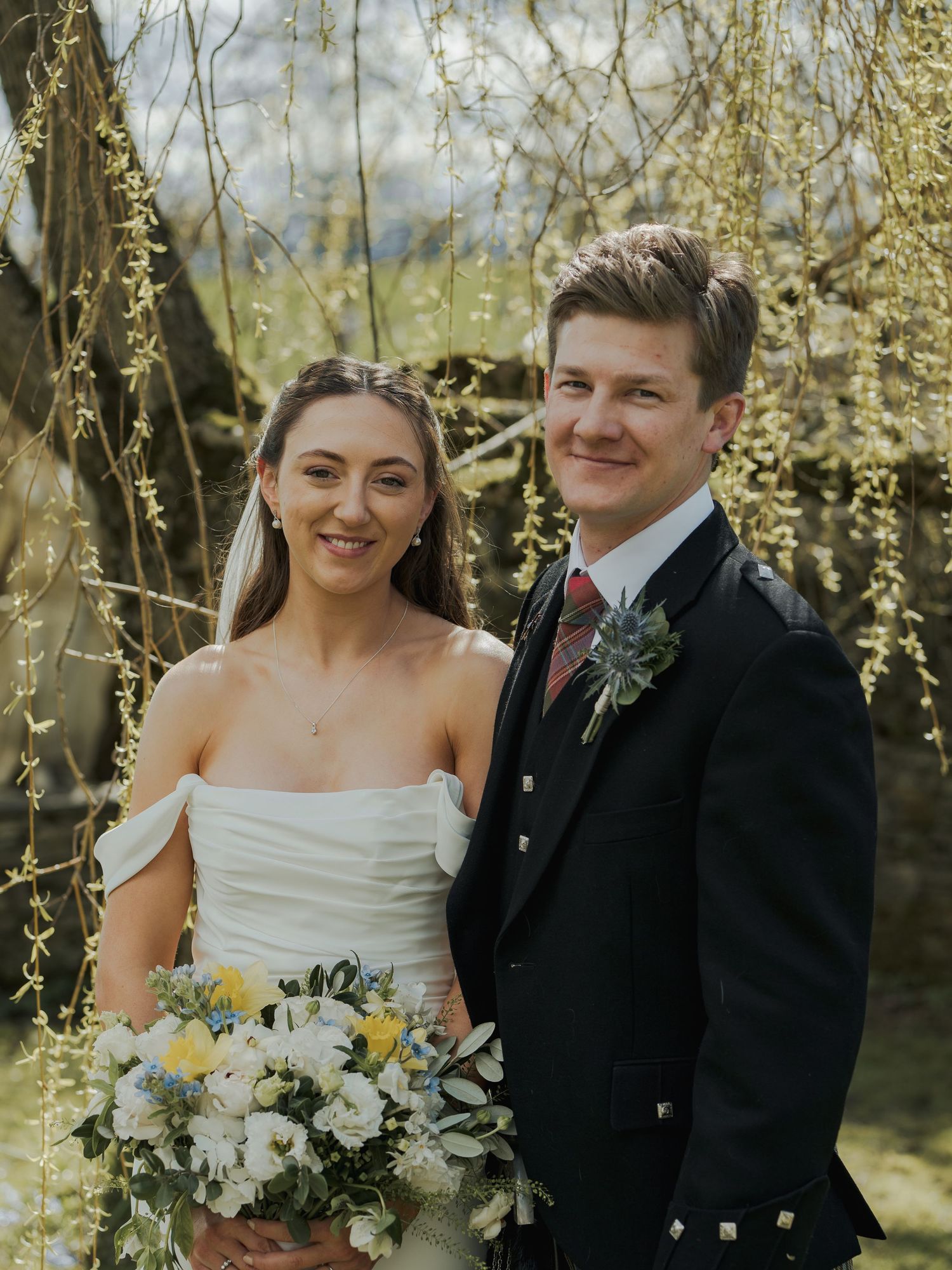 A bride and groom pose outdoors under a weeping willow tree, she holds a white and yellow bouquet.