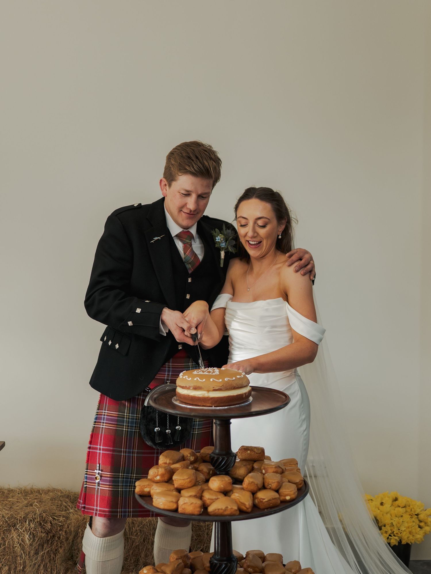 Couple cutting wedding cake in Scottish barn venue, groom in tartan kilt, bride in white off-shoulder dress, profiteroles displayed below.