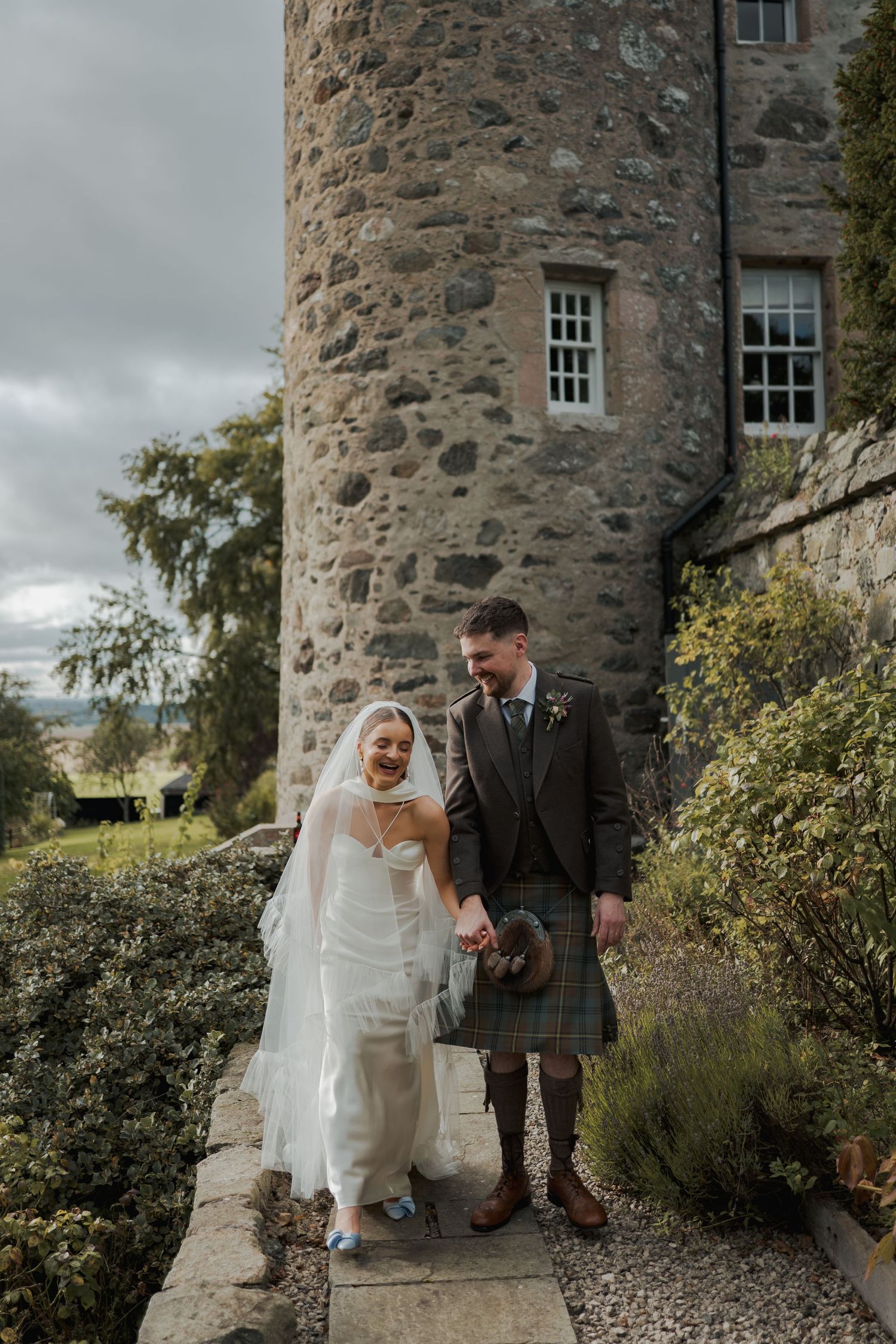 Bride and groom walking hand in hand outside a Scottish castle, groom wearing a tartan kilt, bride in a white gown and veil.