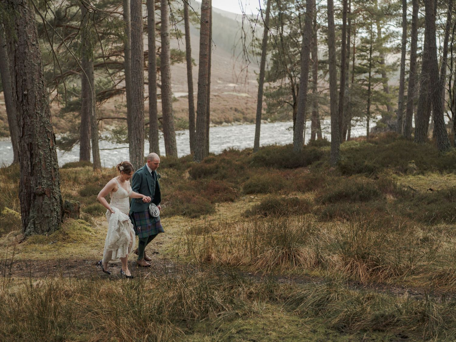 Bride and groom walk hand in hand through a Scottish pine forest with a loch visible in the background.