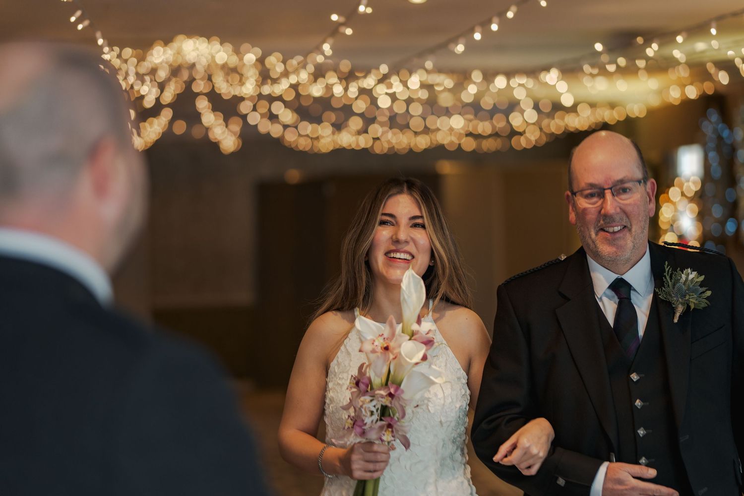 Smiling bride in white halter gown walks down aisle with bouquet, fairy lights glowing overhead at elegant wedding venue.