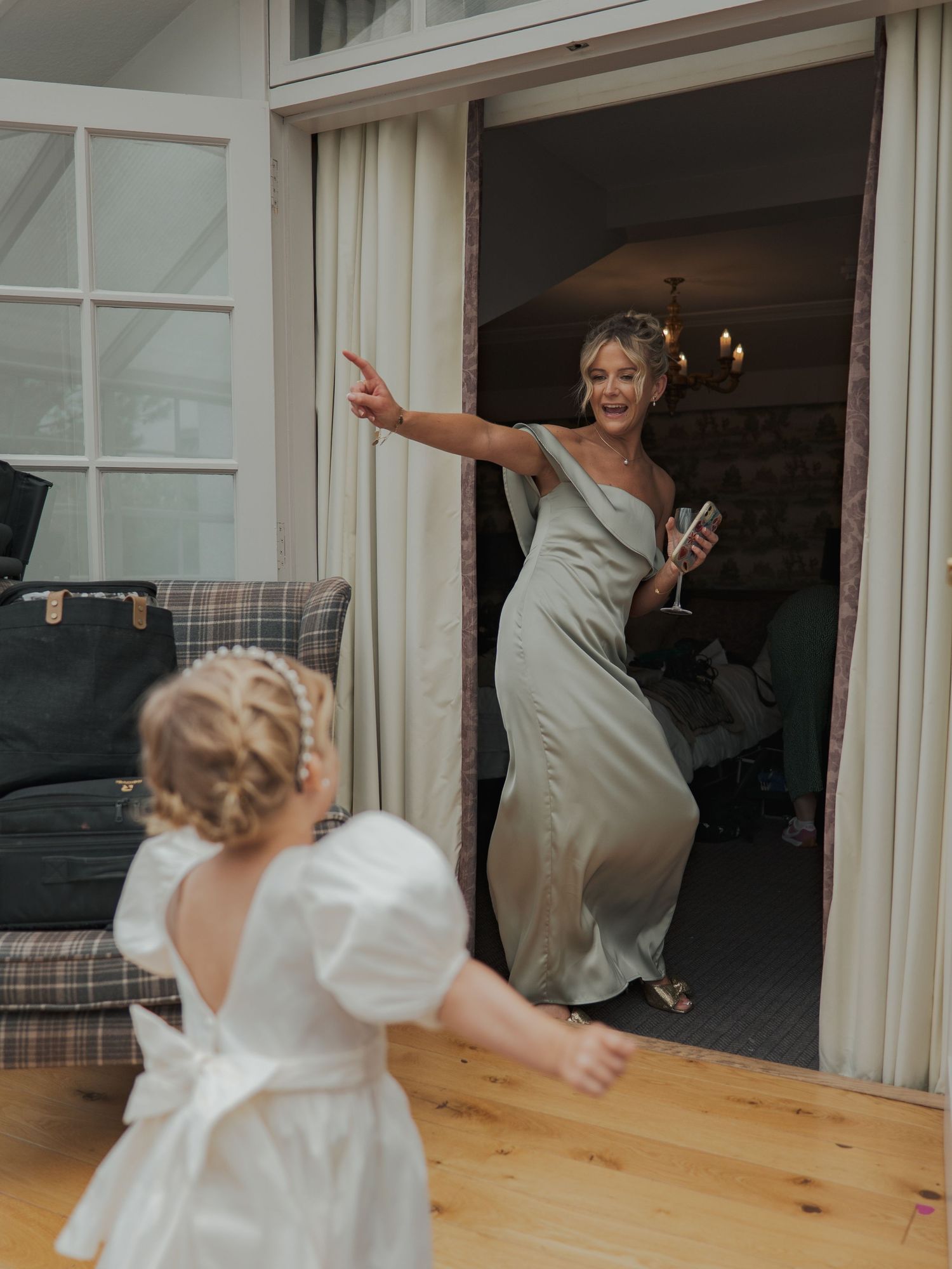 Two people in pale flowing dresses share a joyful dancing moment by large glass doors.