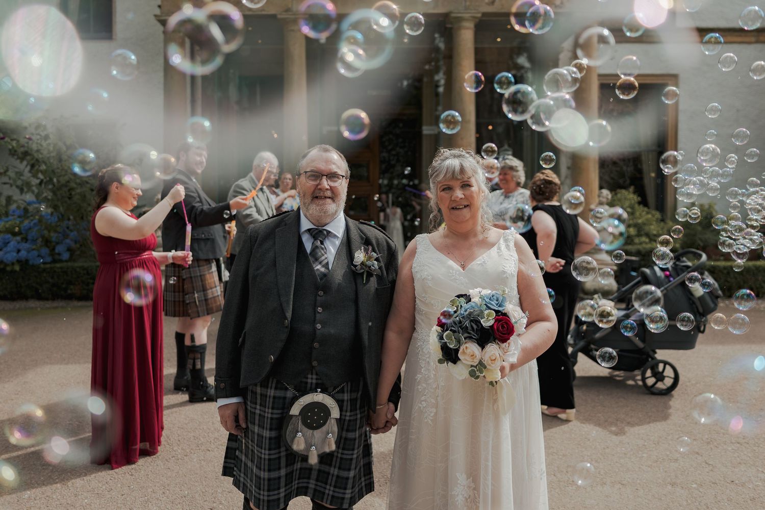 Bride and groom smile amid floating bubbles outside a Scottish wedding venue, groom wearing traditional tartan kilt.