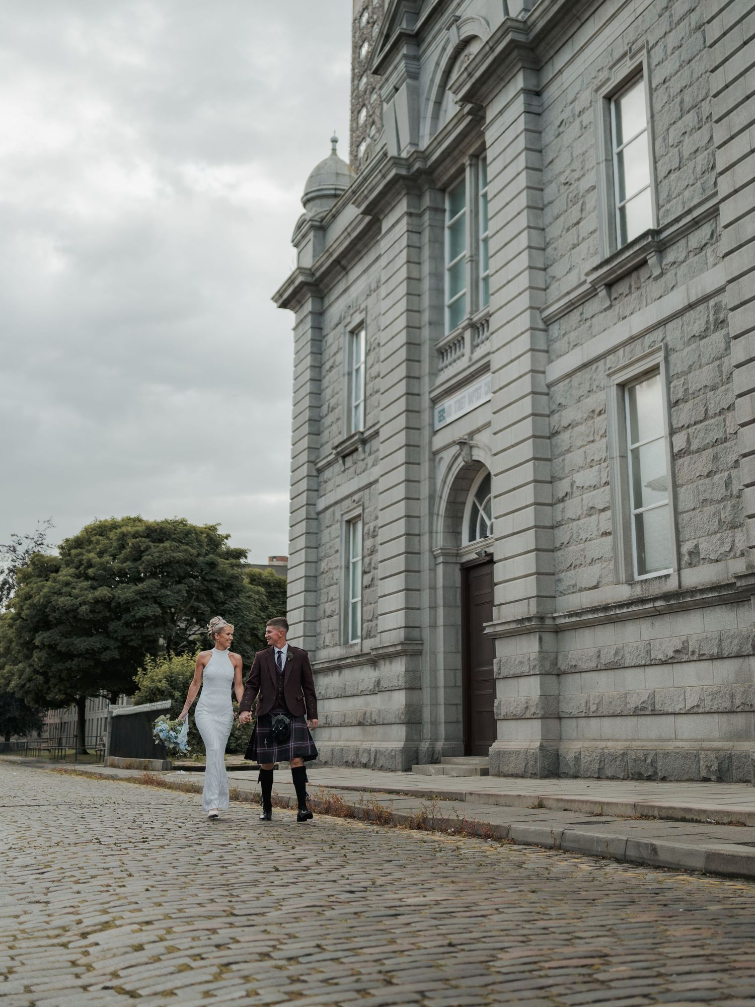 Bride and groom in Scottish kilt walking on cobblestone street beside grand granite church on cloudy day.