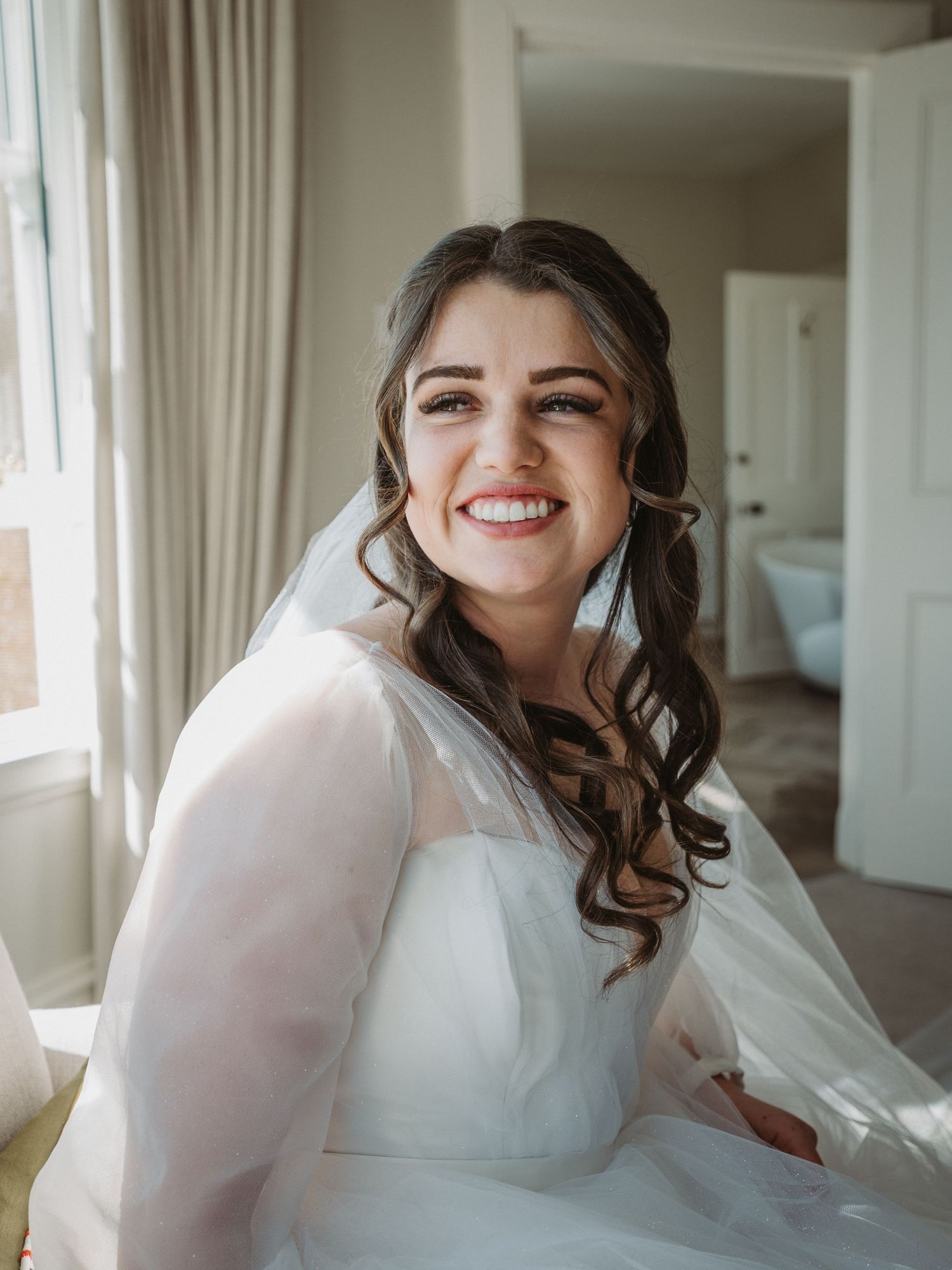 A bride in a white wedding dress with sheer sleeves smiles joyfully by a sunlit window in Kinclune House.