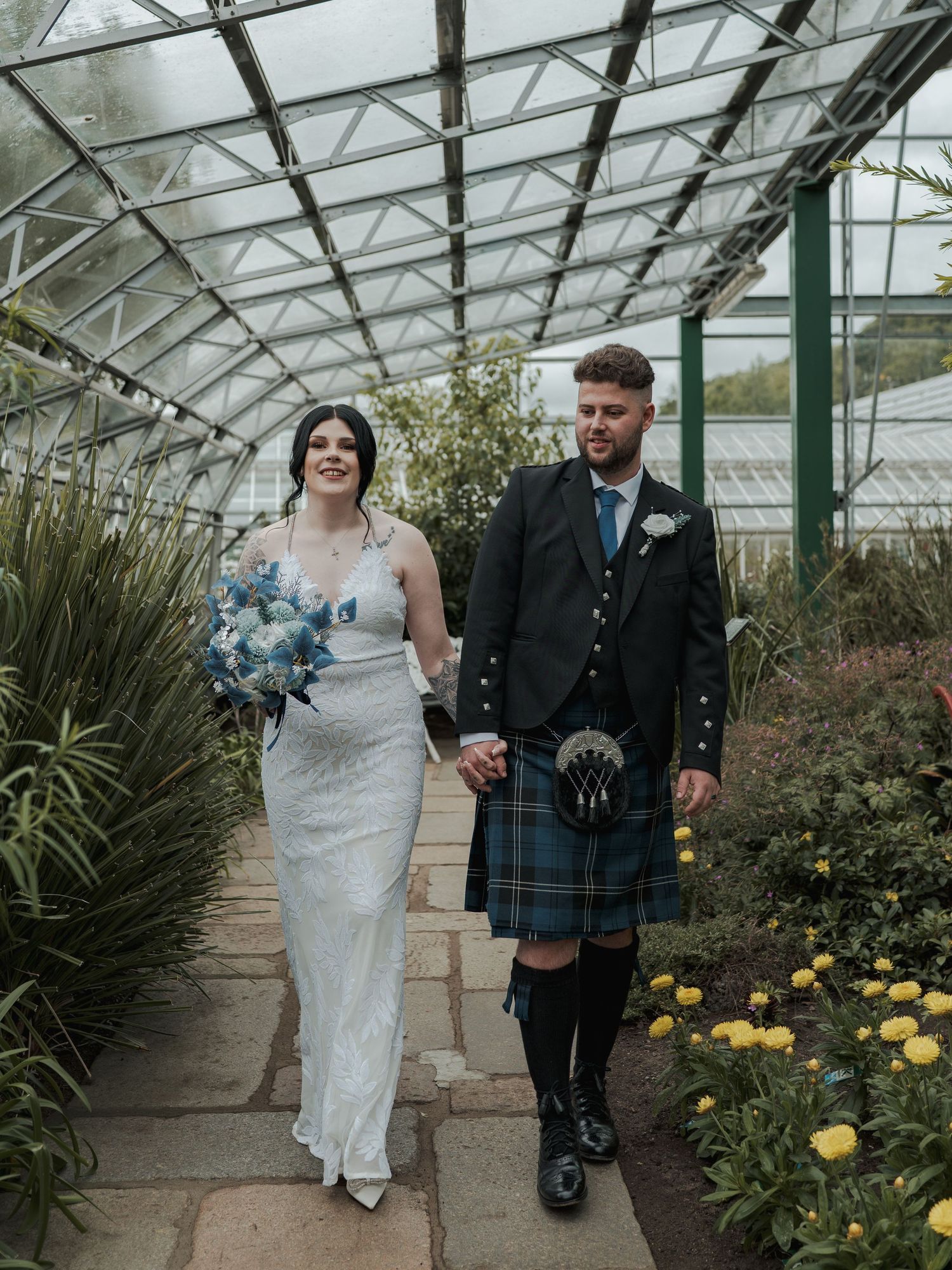 A bride and groom walk hand in hand through a greenhouse garden, she in a white gown, he in a Scottish tartan kilt.
