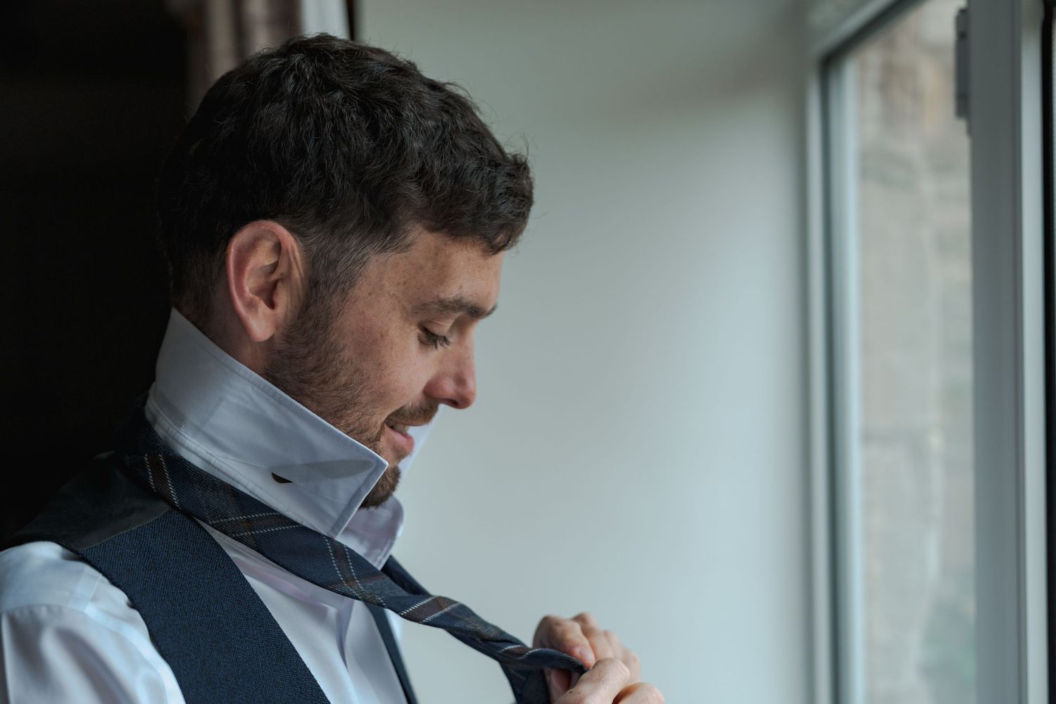 A person wearing a vest and collared shirt adjusts their tie while looking thoughtfully out a window.