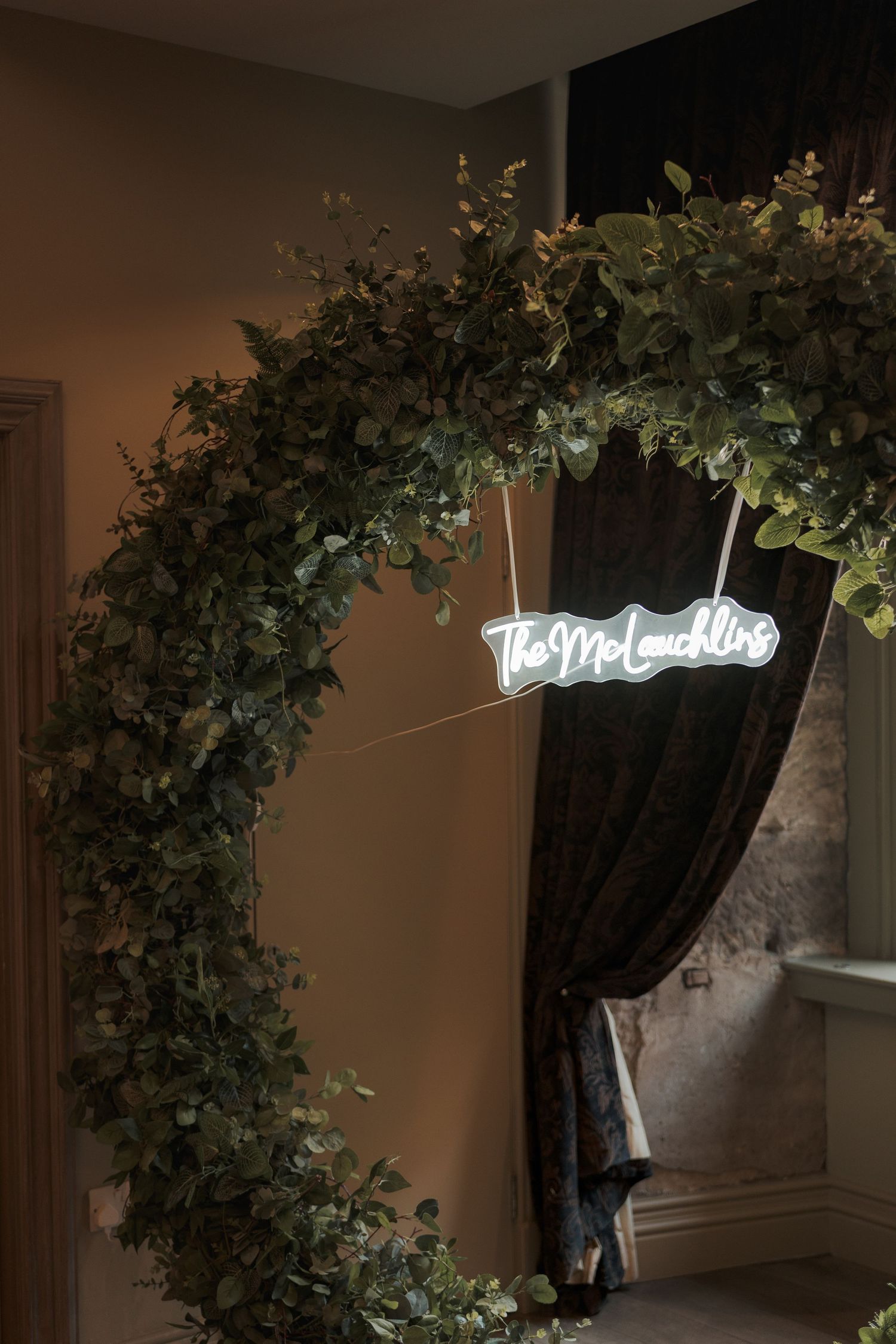 A decorative greenery arch with a glowing neon 'welcome' sign hangs over an elegant curtained doorway.