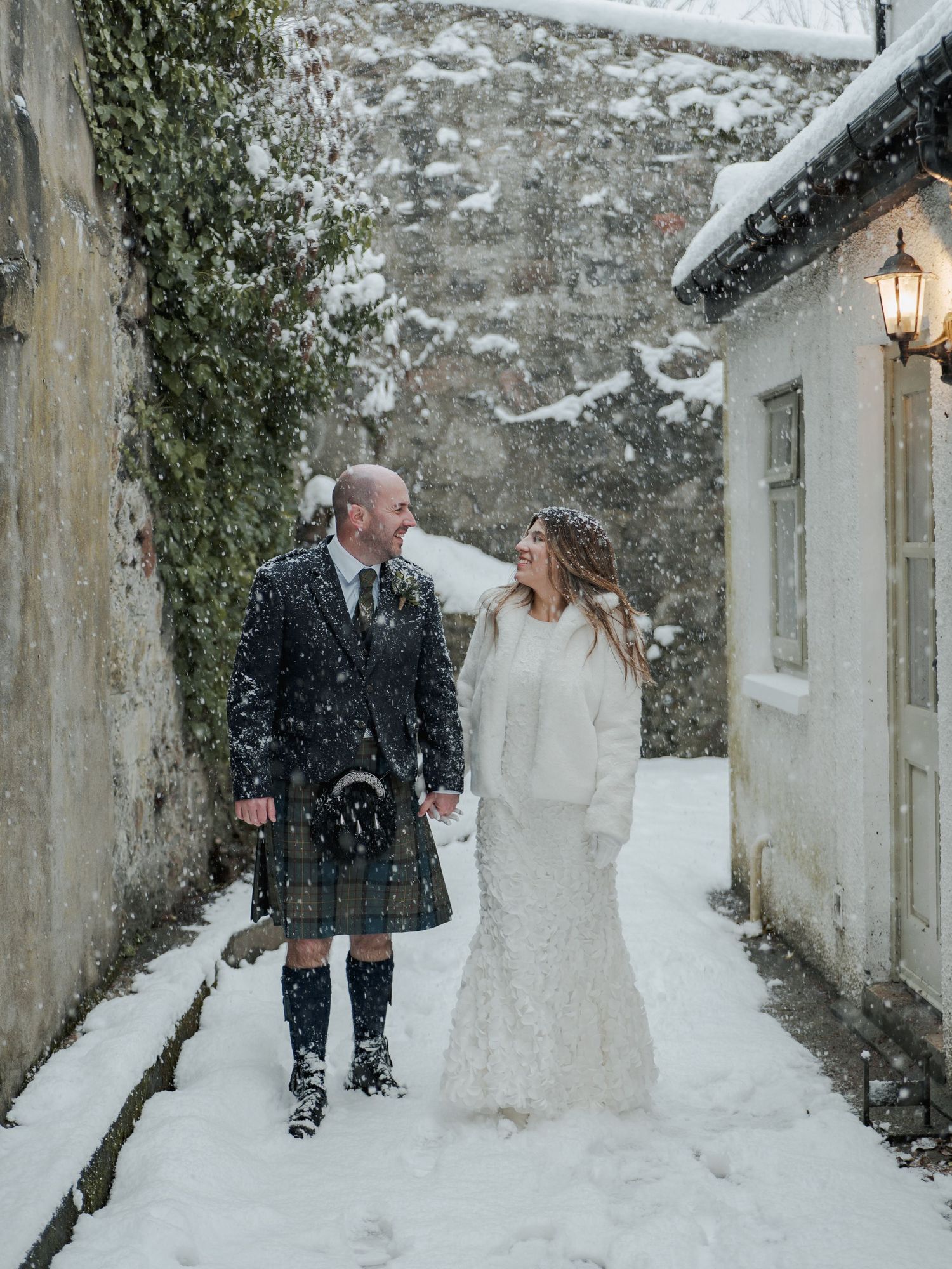 A couple in wedding attire walks along a snowy stone path with lanterns illuminating the winter scene.