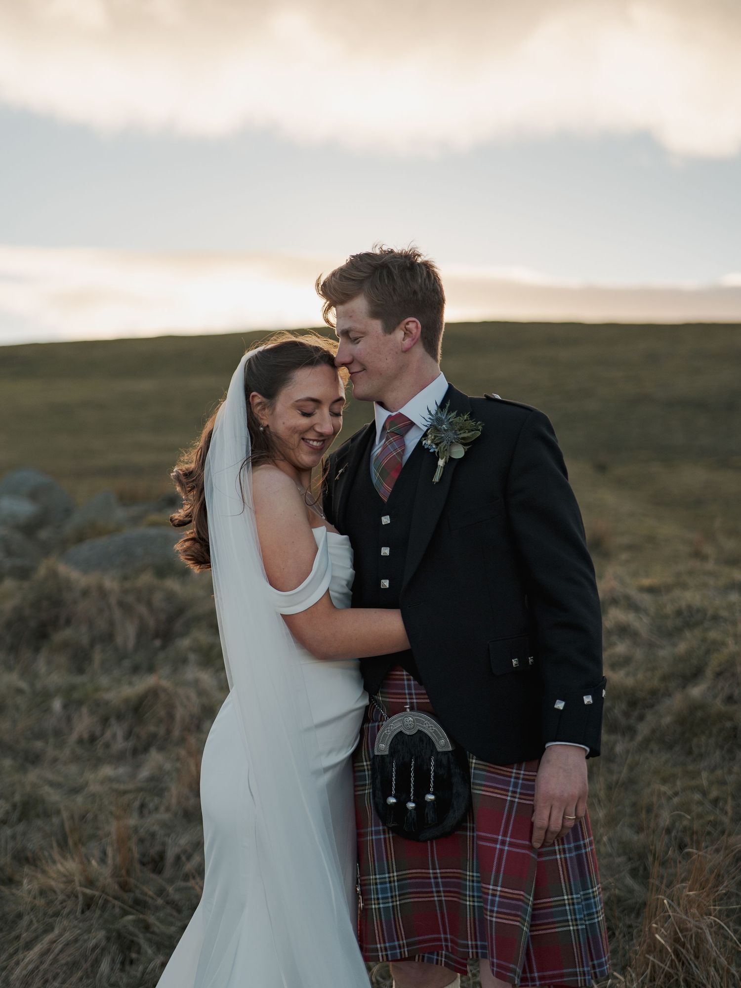 A bride and groom embrace in a Scottish highland landscape, the groom wearing a traditional tartan kilt at sunset.