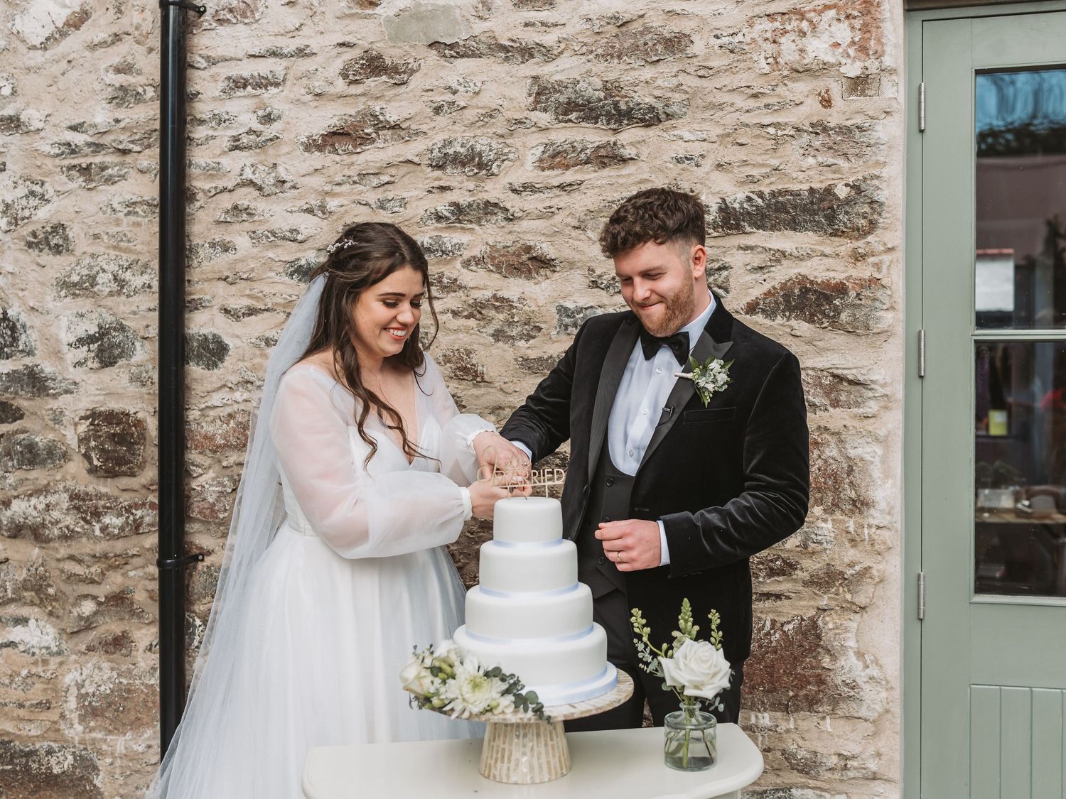 Newlyweds cutting their elegant white tiered wedding cake together against stone wall backdrop of Kinclune Steading.