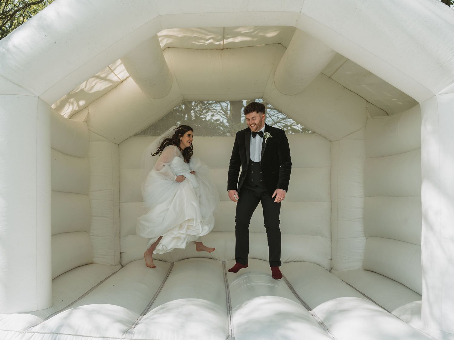 A couple in wedding attire jumps playfully inside a white inflatable bounce house.