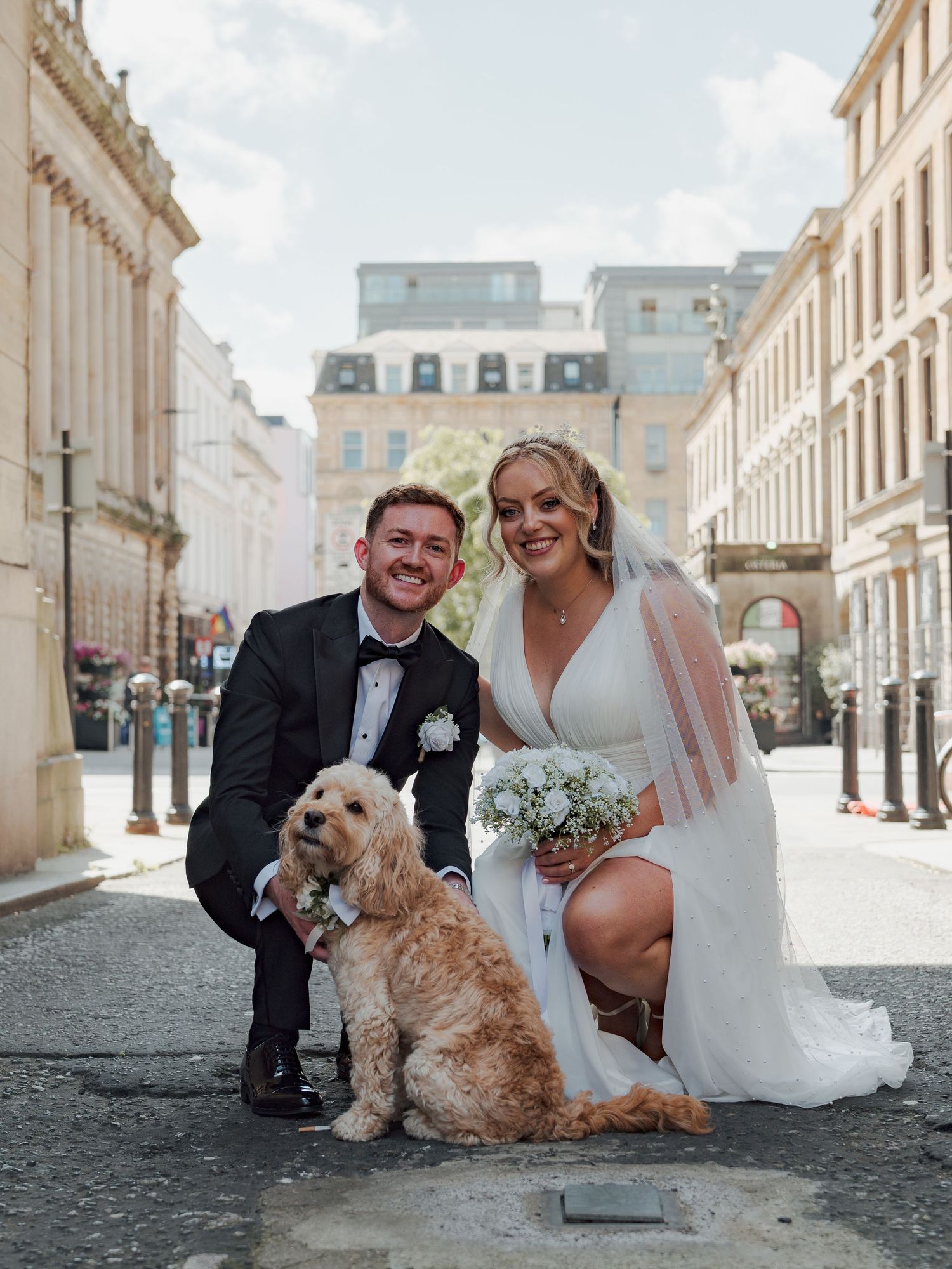 A wedding couple poses with their small brown dog on a cobblestone street in front of historic Glasgow buildings.
