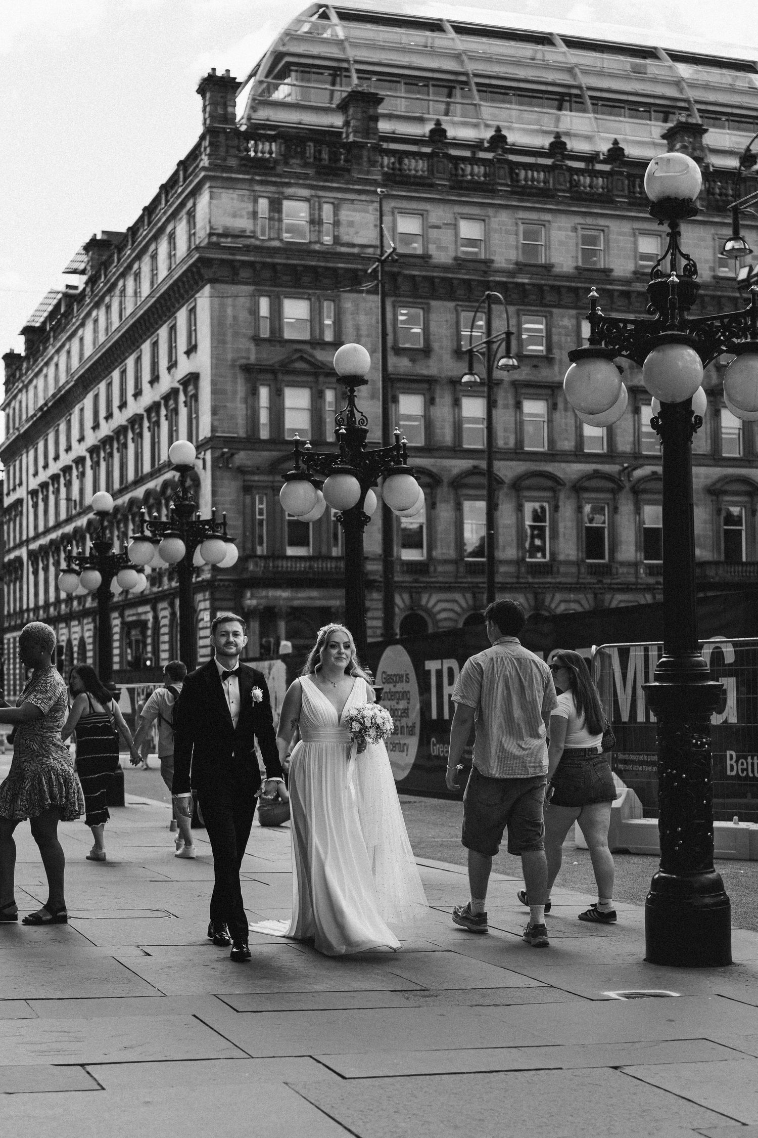 Newlyweds walk down a city street with ornate historic architecture and vintage lamp posts while passersby stroll by.