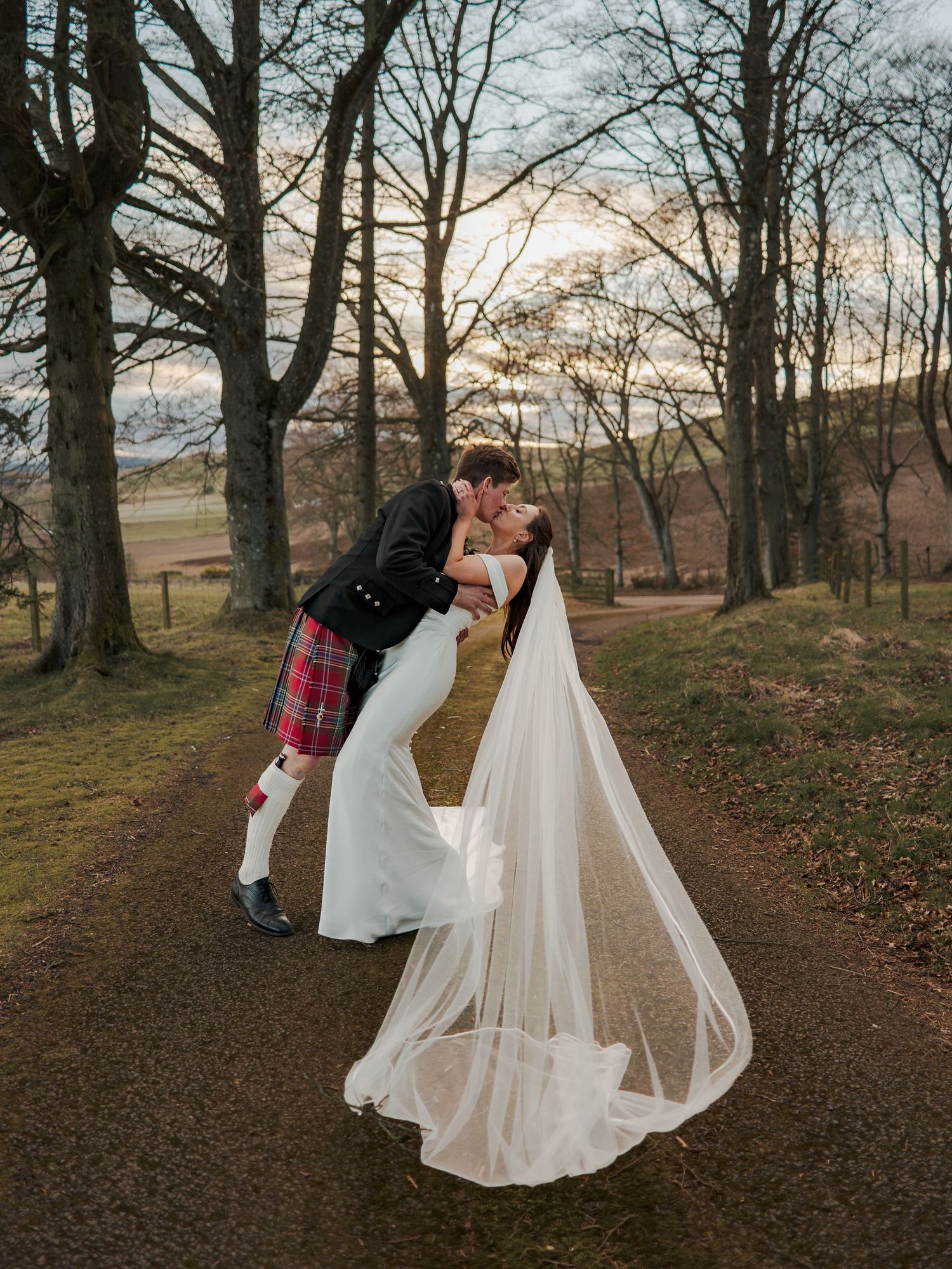 Bride and groom share a romantic dip kiss on a tree-lined path, groom in red tartan kilt, bride in flowing white gown with veil.