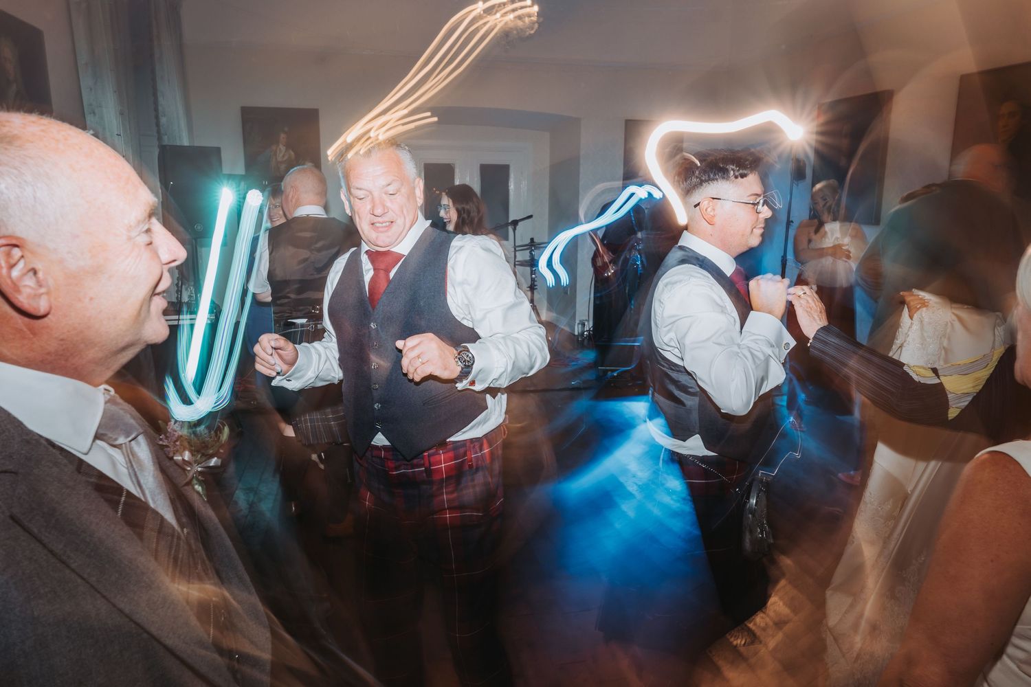Wedding reception guests dancing in dark room with blue neon lights and traditional Scottish kilts.