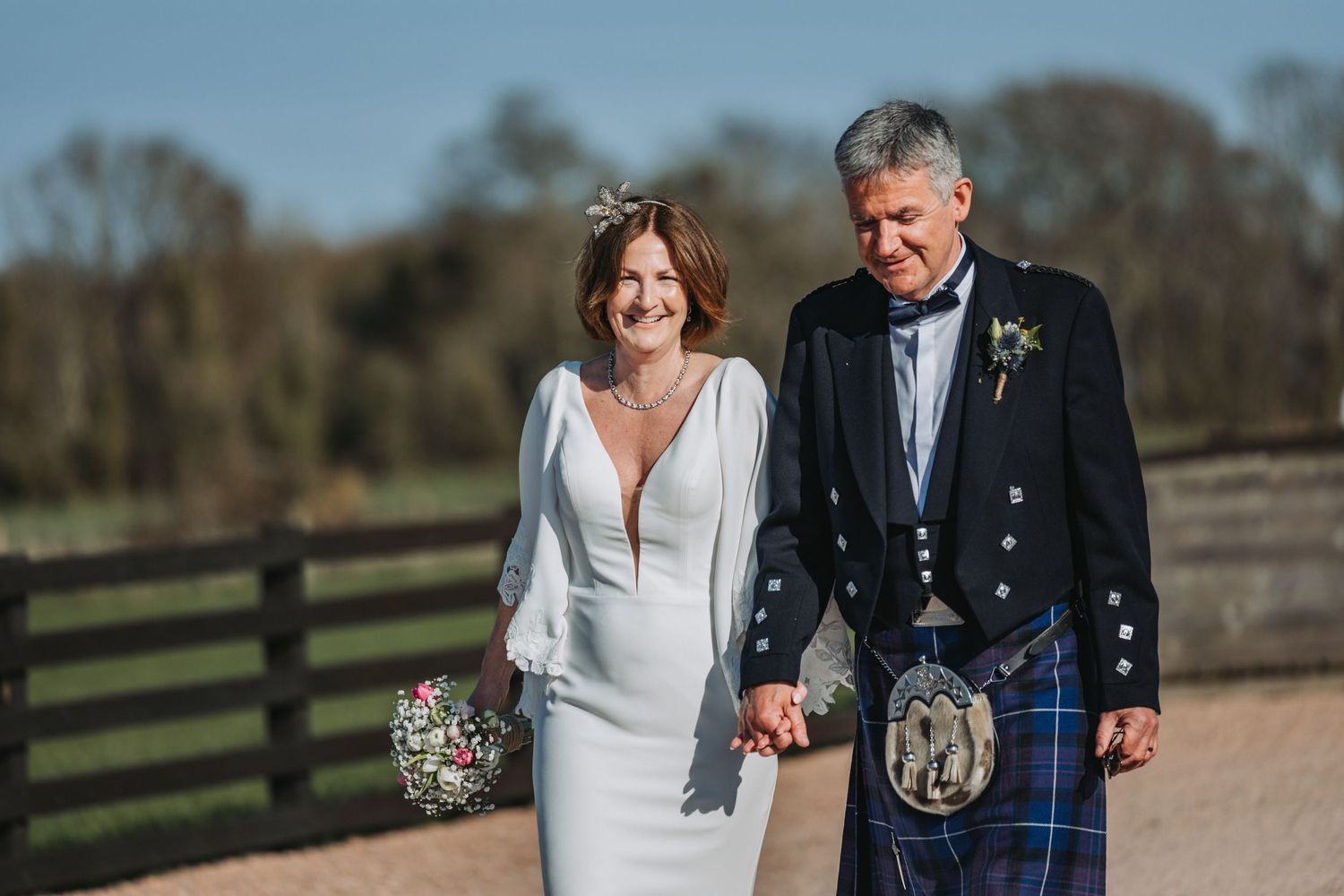 Couple walking hand in hand outdoors at Falside Mill, groom in Scottish kilt and bride in elegant white dress.