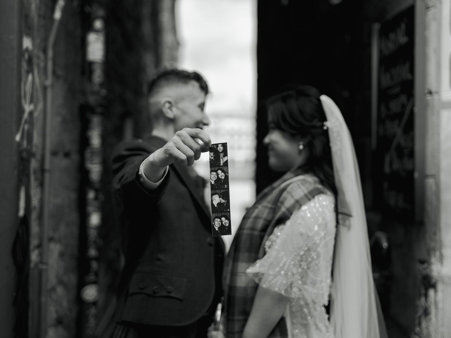 Black and white photo of a groom showing photo booth strip to smiling bride in wedding attire outdoors in Edinburgh