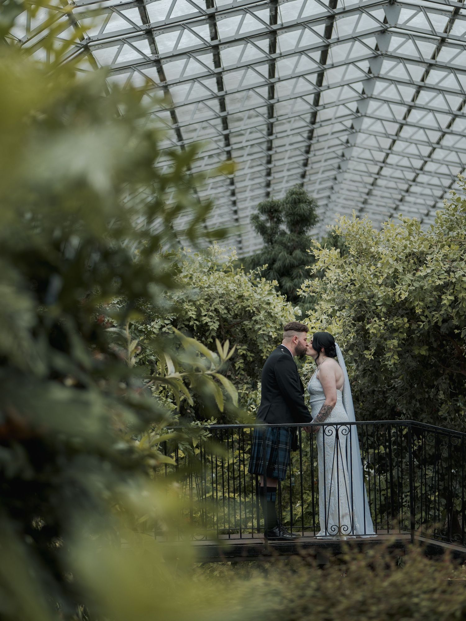 Bride and groom share a romantic moment on a balcony inside a stunning glass greenhouse surrounded by lush greenery.