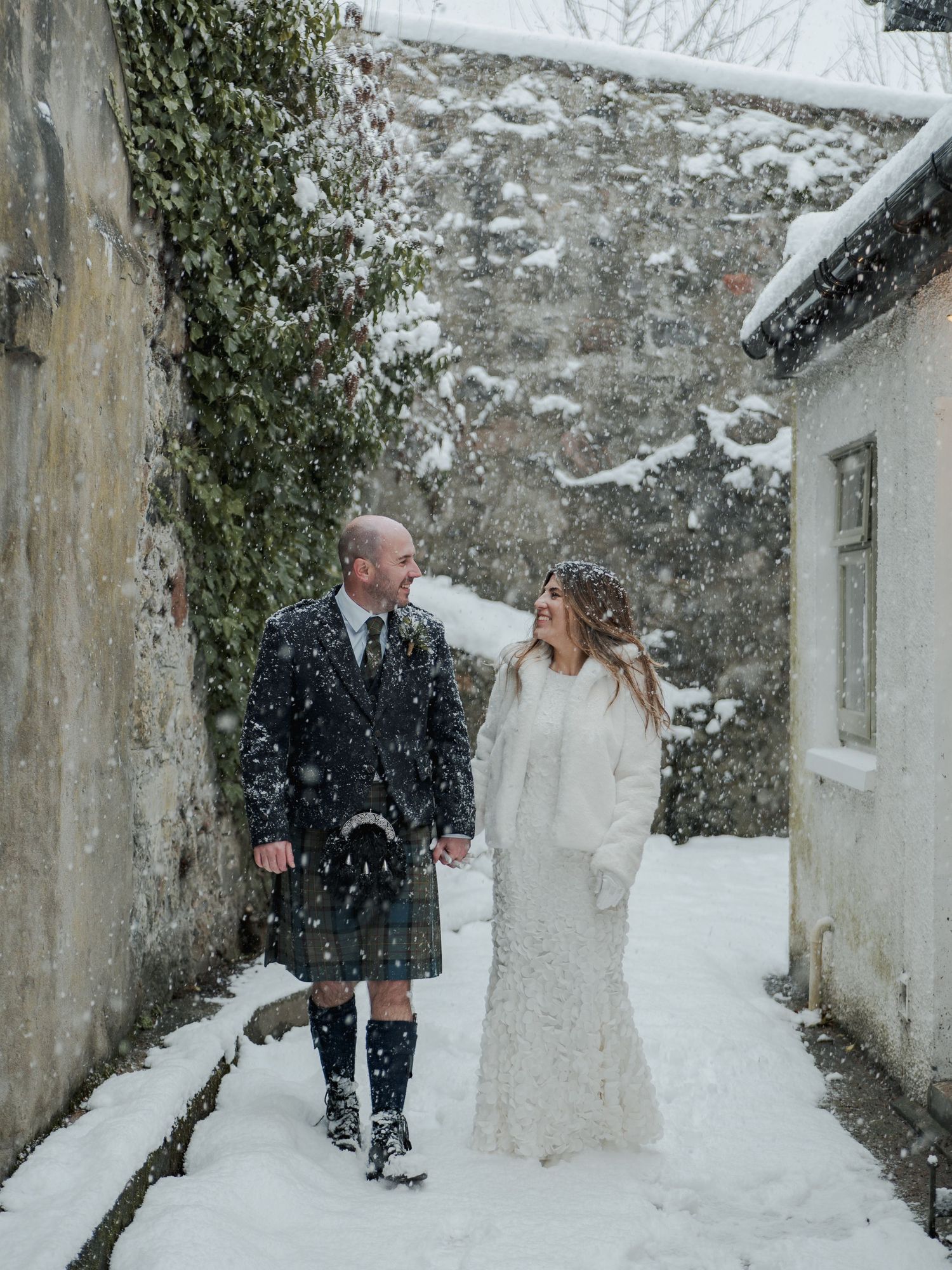 Couple walking in snowy courtyard, groom in Scottish kilt, bride in white lace gown with fur wrap.