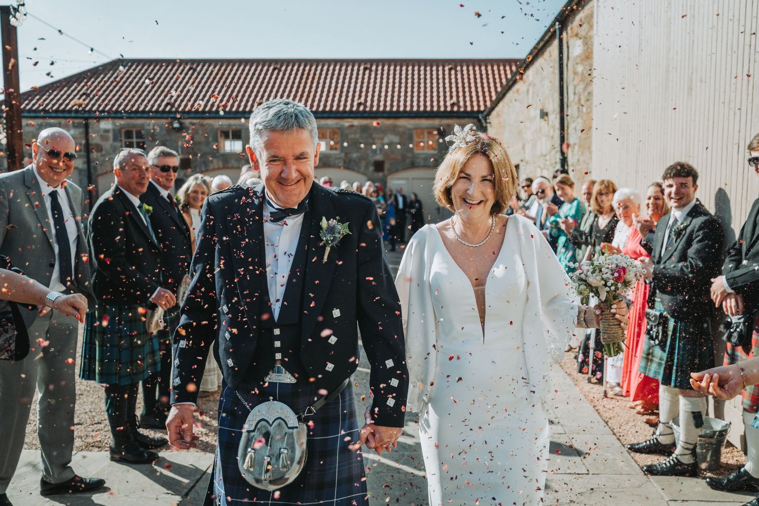 Happy couple exiting Falside Mill while guests throw confetti, groom wearing traditional Scottish kilt.