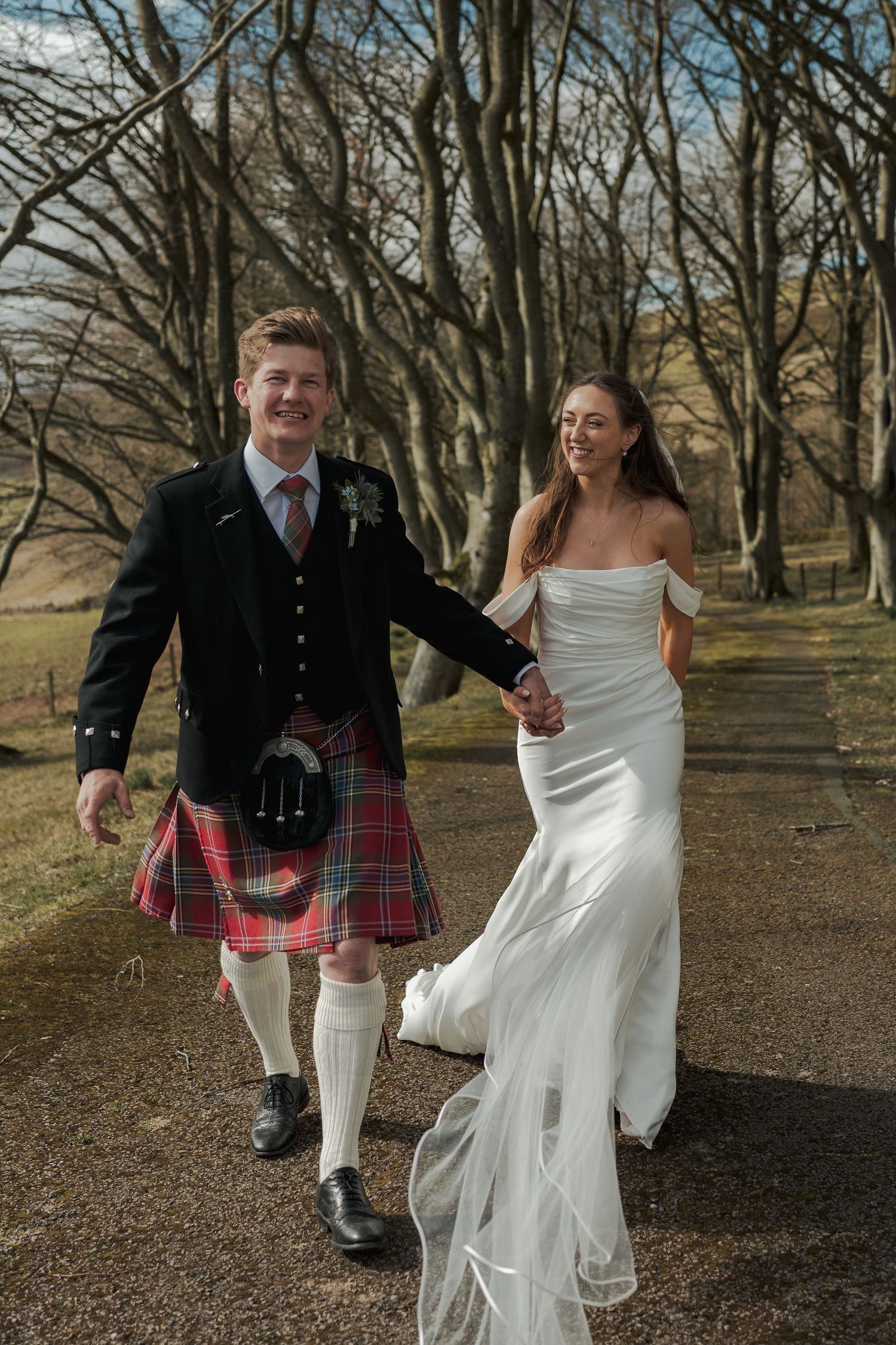 Happy newlyweds walk hand-in-hand, groom in Scottish kilt and bride in white off-shoulder gown, among bare winter trees.