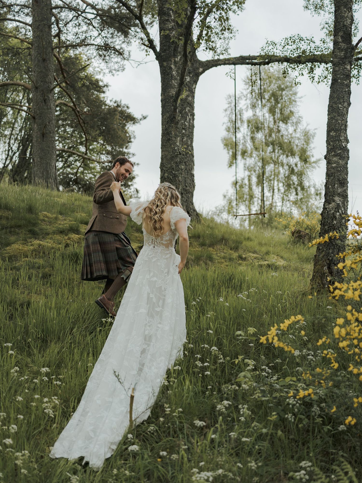 A bride in lace gown and groom in tartan kilt dance together in a lush Scottish woodland meadow.