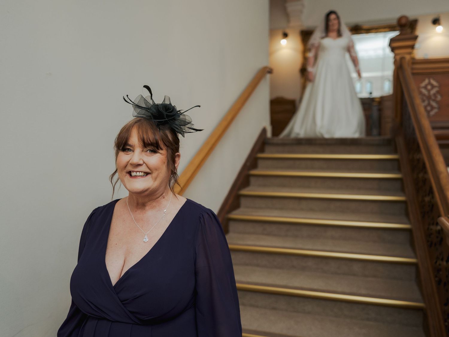 A smiling woman in navy dress with fascinator stands before a bride descending an elegant wooden staircase.