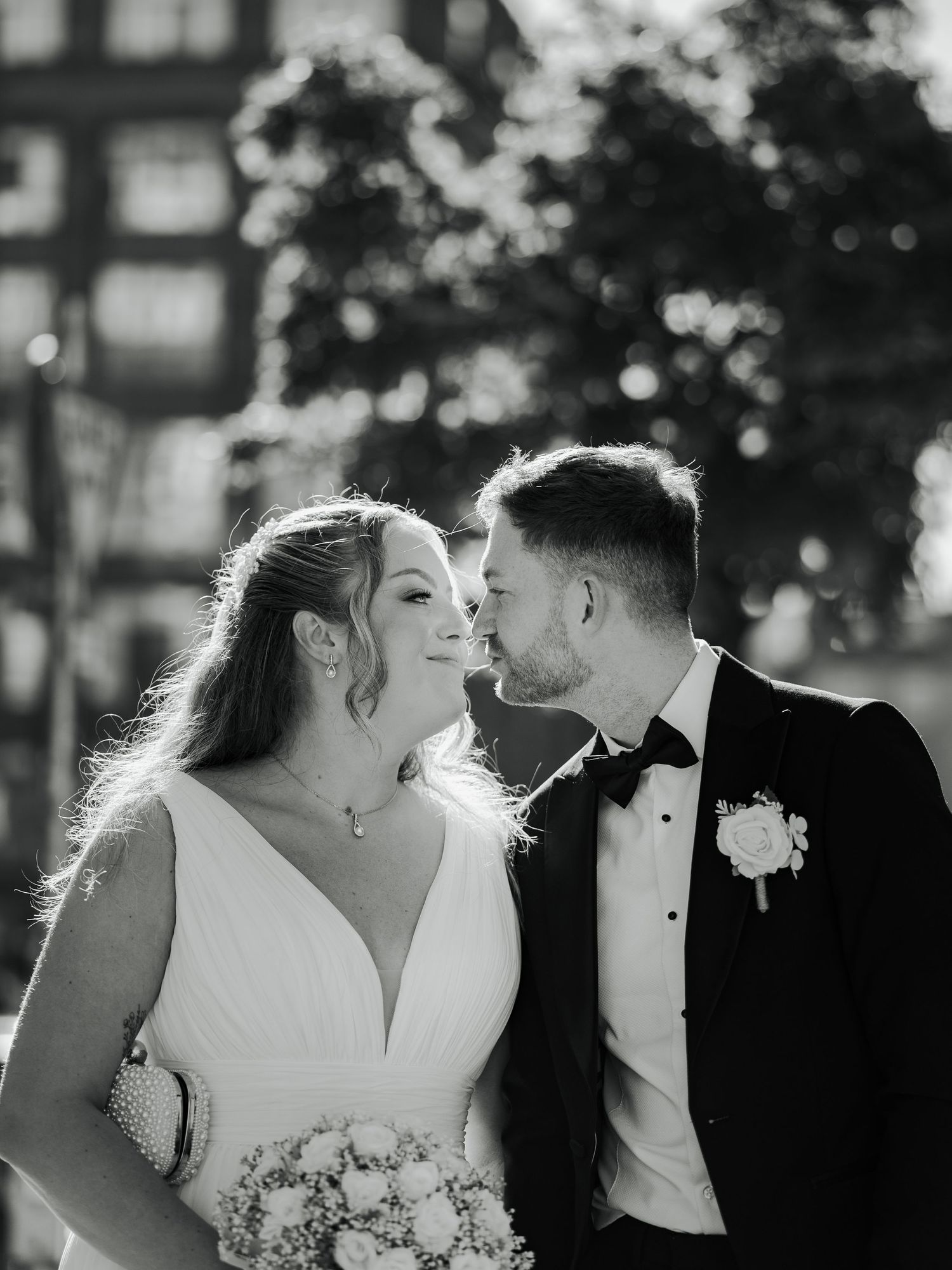 Black and white portrait of newlyweds sharing an intimate moment outdoors with sunlight filtering through trees.