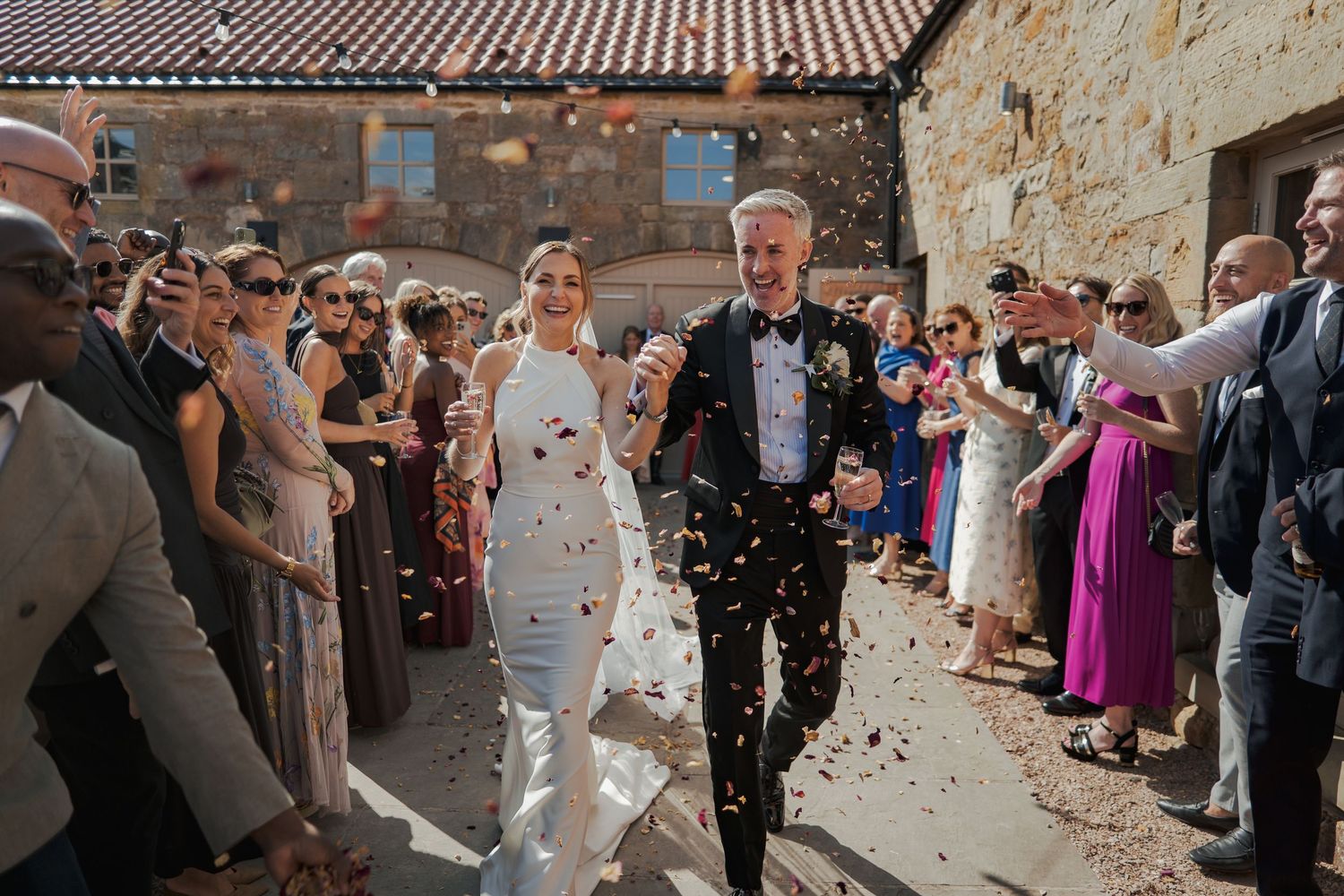 Bride and groom walk through confetti shower surrounded by cheering wedding guests outside rustic stone venue.