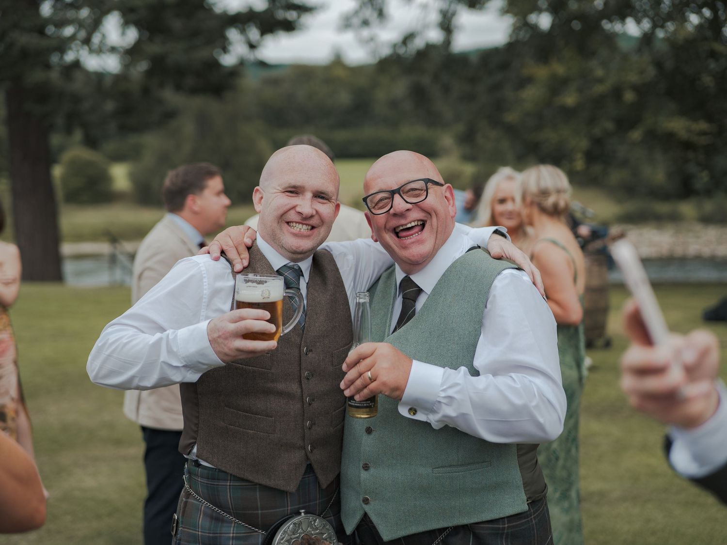 A group of wedding guests enjoying drinks and laughter in an outdoor setting with trees in the background.