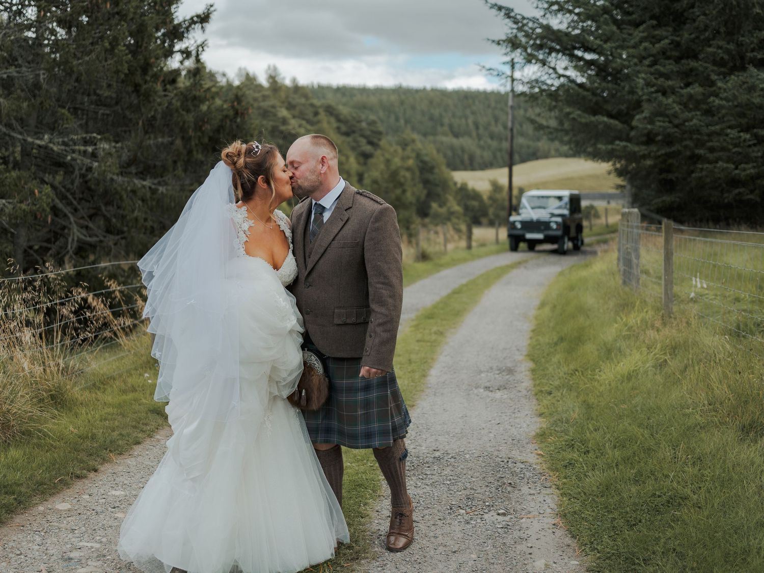 Bride and groom share a romantic moment on a Scottish country path, groom wearing traditional tartan kilt.