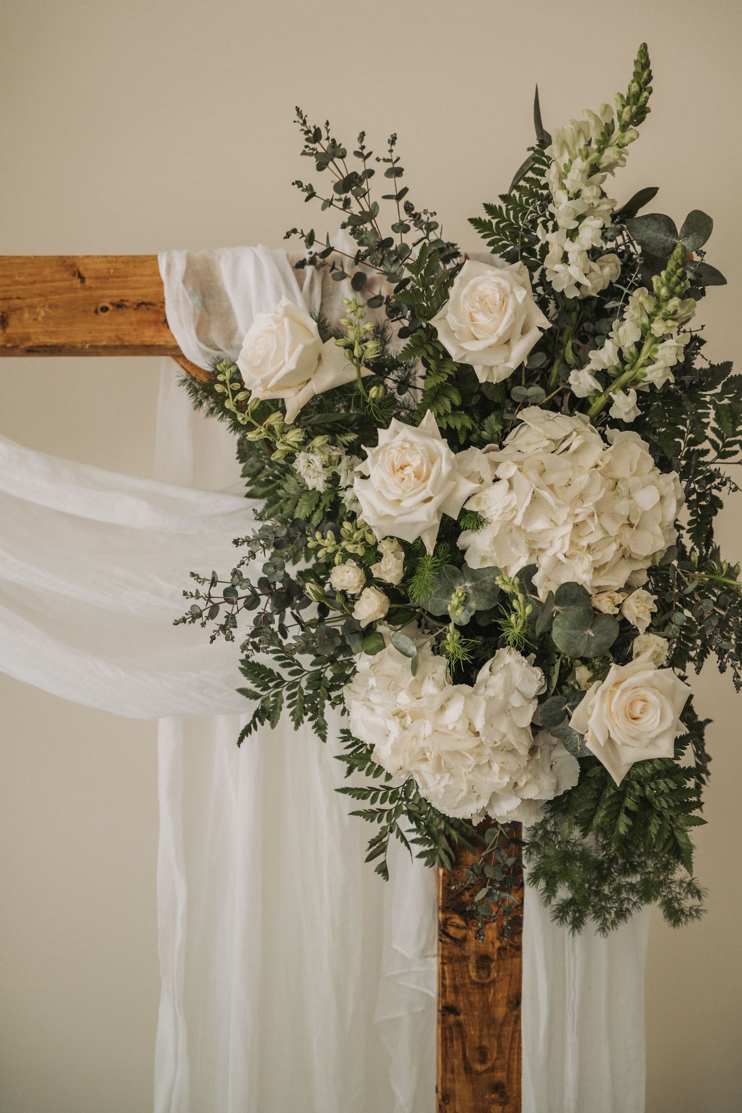 White roses and hydrangeas with green eucalyptus arranged in rustic wooden decor with flowing white fabric backdrop.