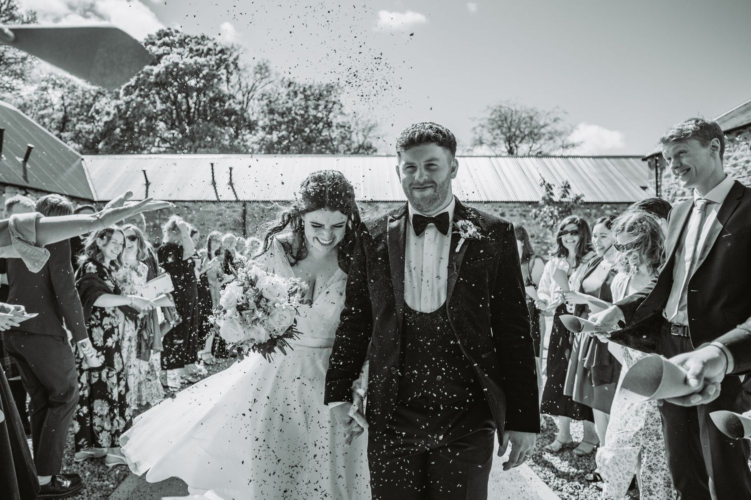 Wedding guests celebrate by throwing rice and confetti at an outdoor celebration under a clear sky.