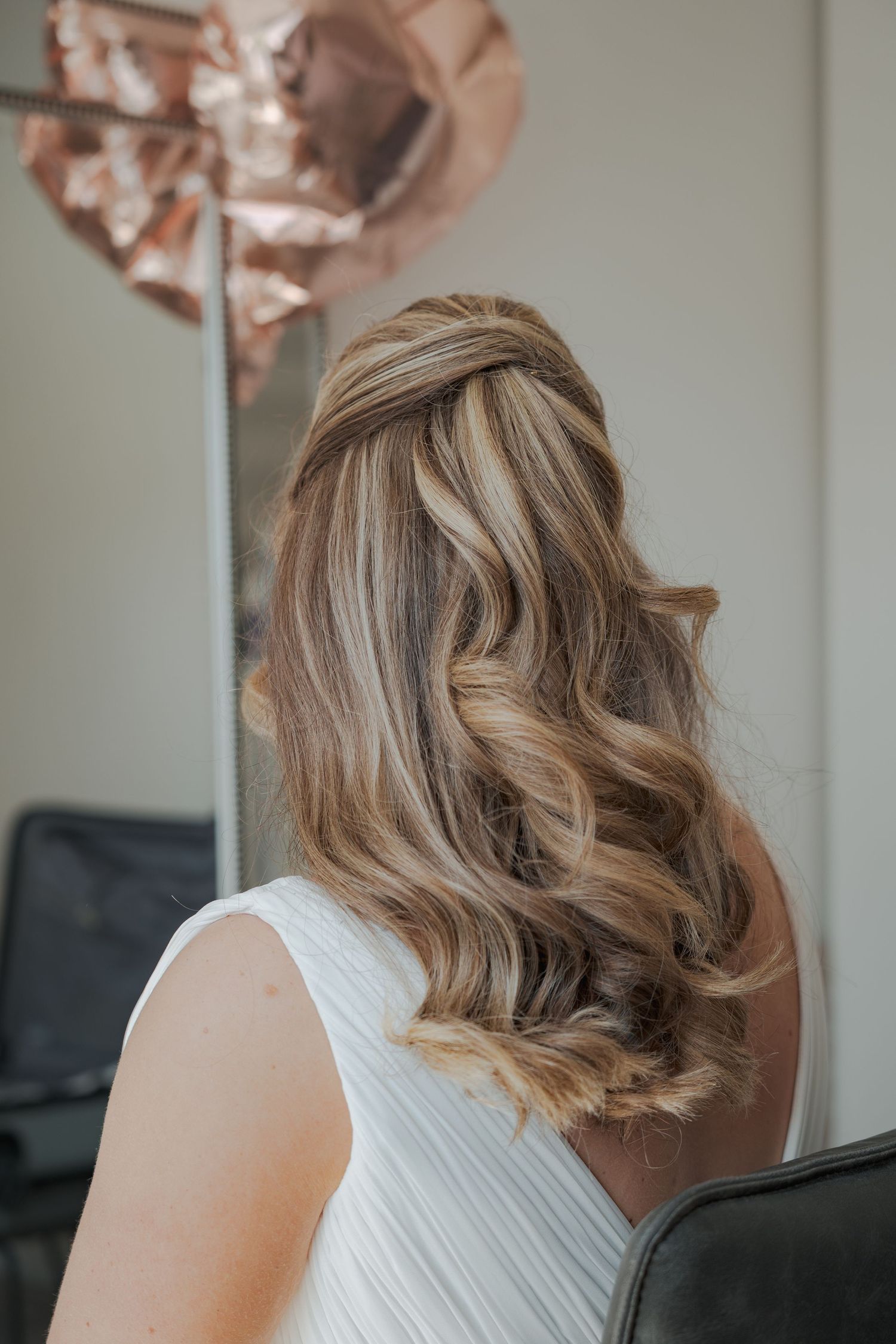 Elegant blonde wavy hairstyle styled in soft curls with a rose gold balloon decoration in the background.