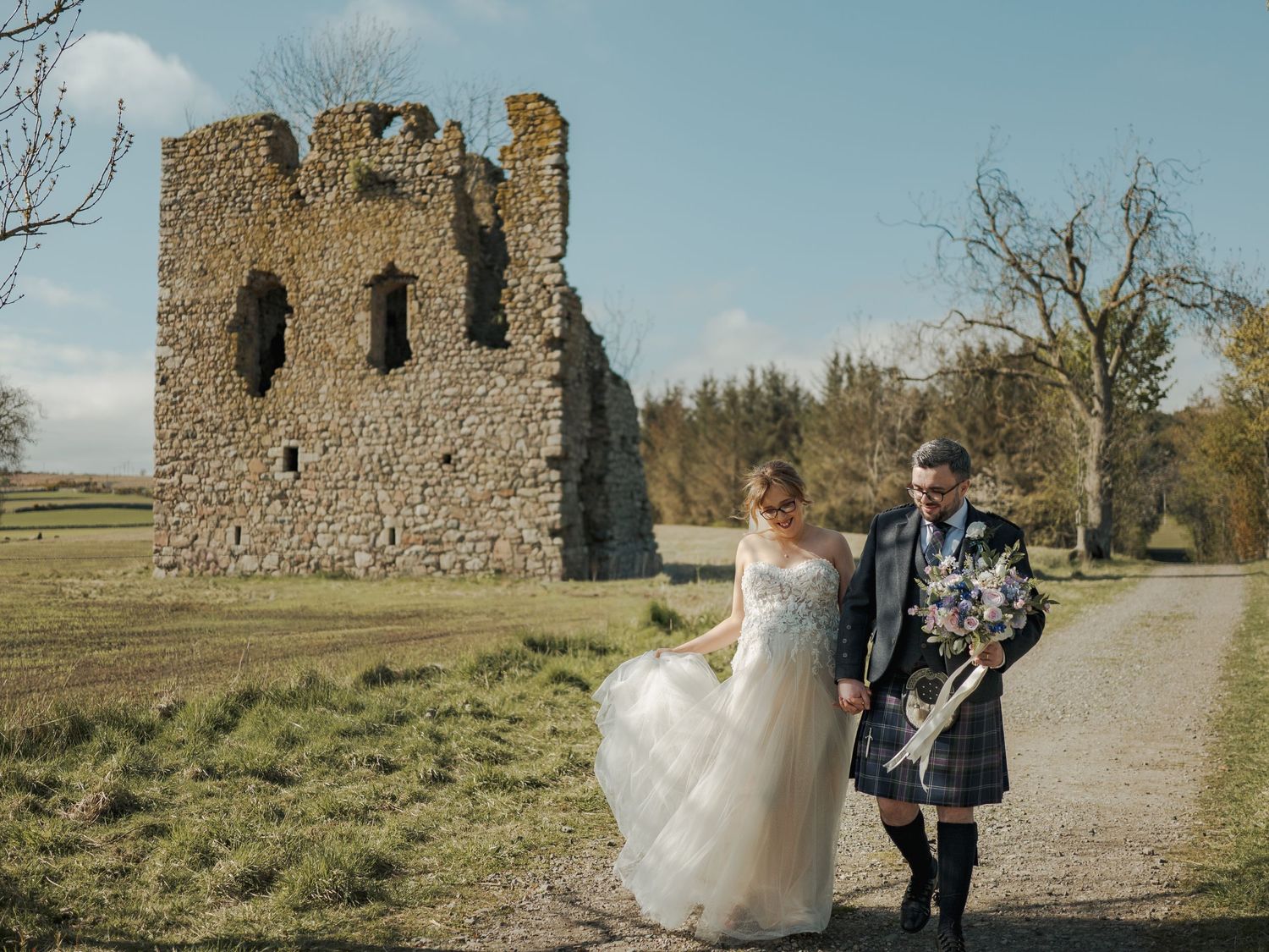 Bride and groom walking near ancient castle ruins in Scotland, groom wearing traditional kilt, bride in elegant white gown.
