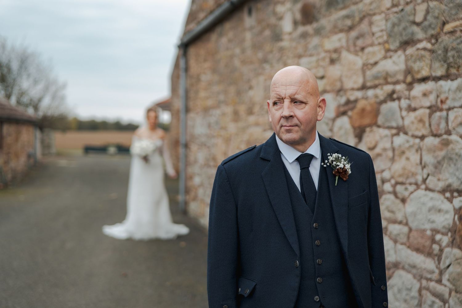 A wedding couple and groomsmen in black suits stand in front of a rustic stone wall on an overcast day.