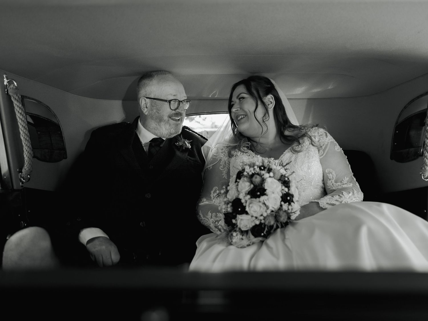 Black and white photo of a bride in a lace gown and older man in a suit sharing a joyful moment inside a wedding car.