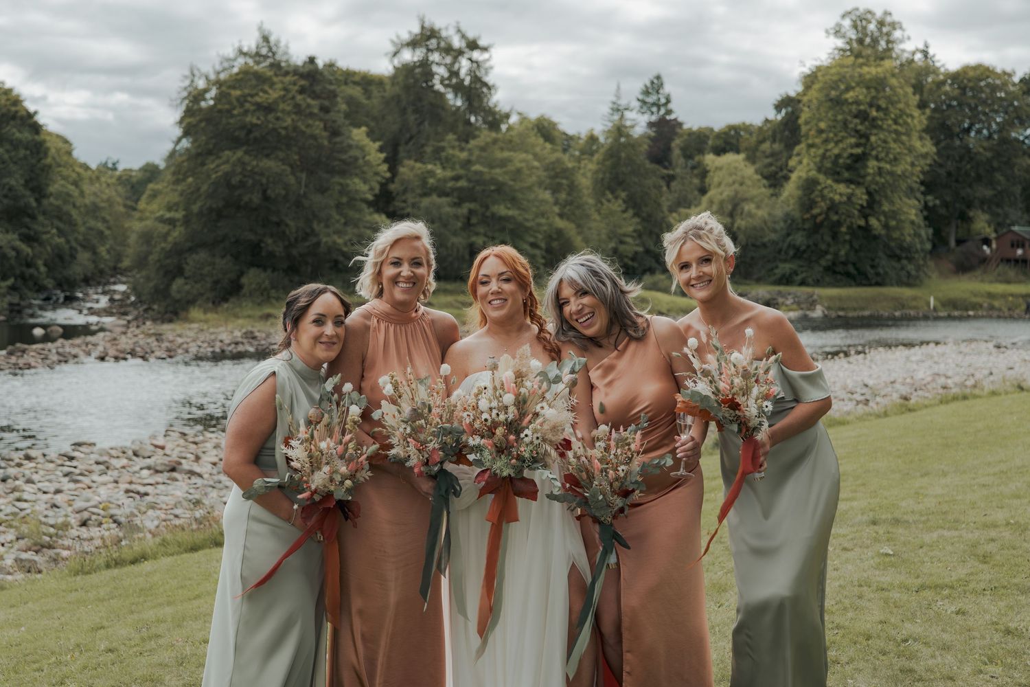 Wedding party stands together holding bouquets of autumn flowers in blush pink and rust colored dresses.
