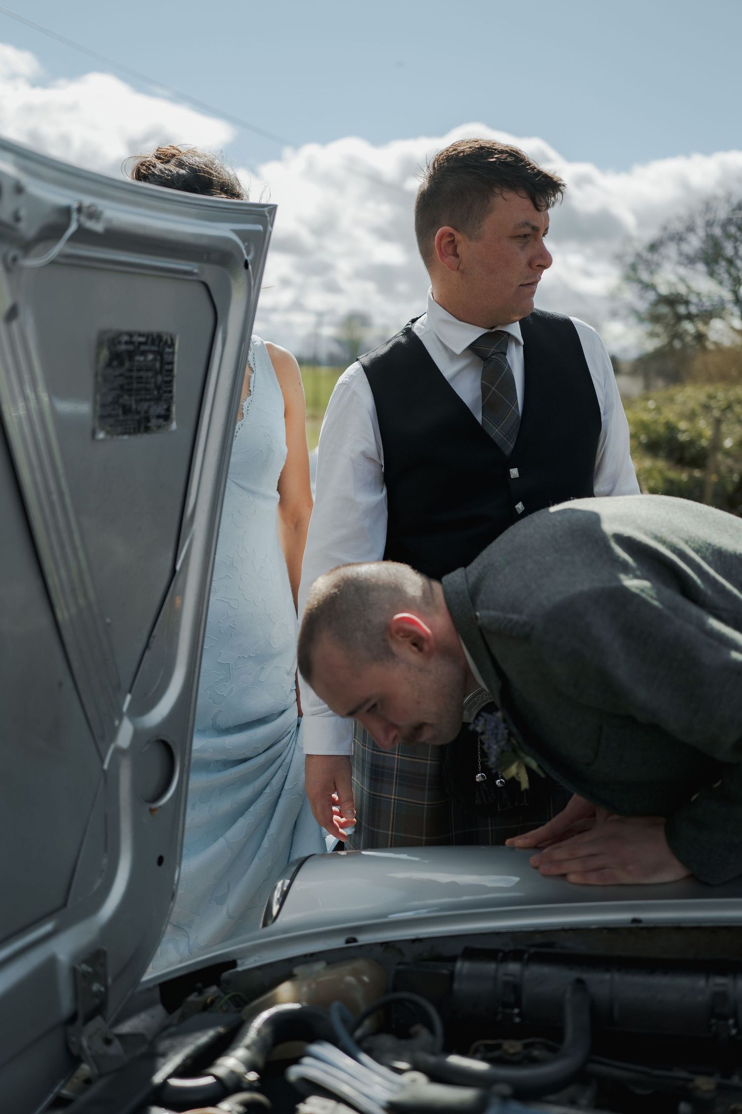 Two men in formal attire inspect a car engine with hood open on a sunny day outdoors.