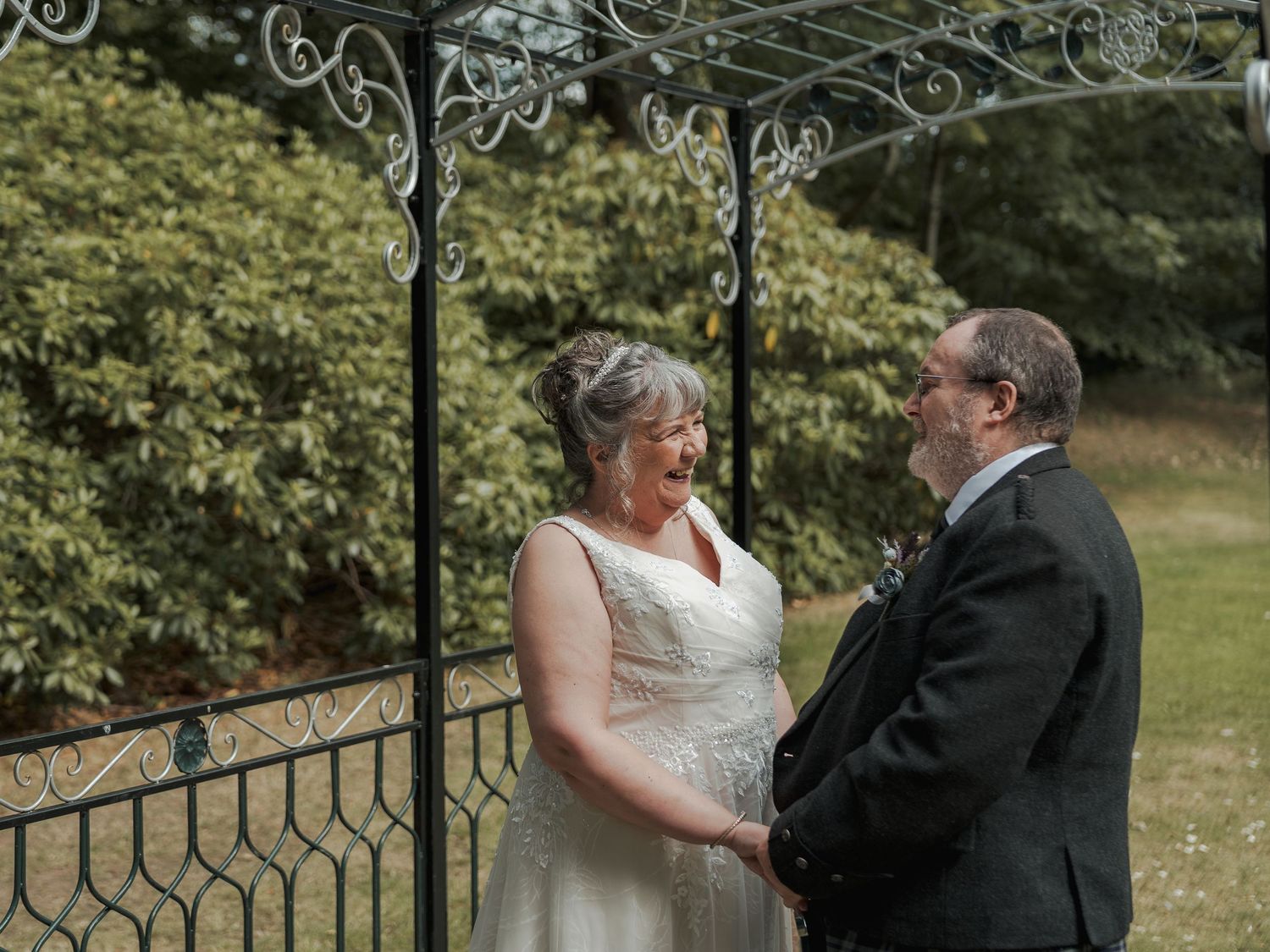 Couple exchanging vows outdoors under decorative iron arbor, bride in white gown laughing joyfully with groom in dark suit.